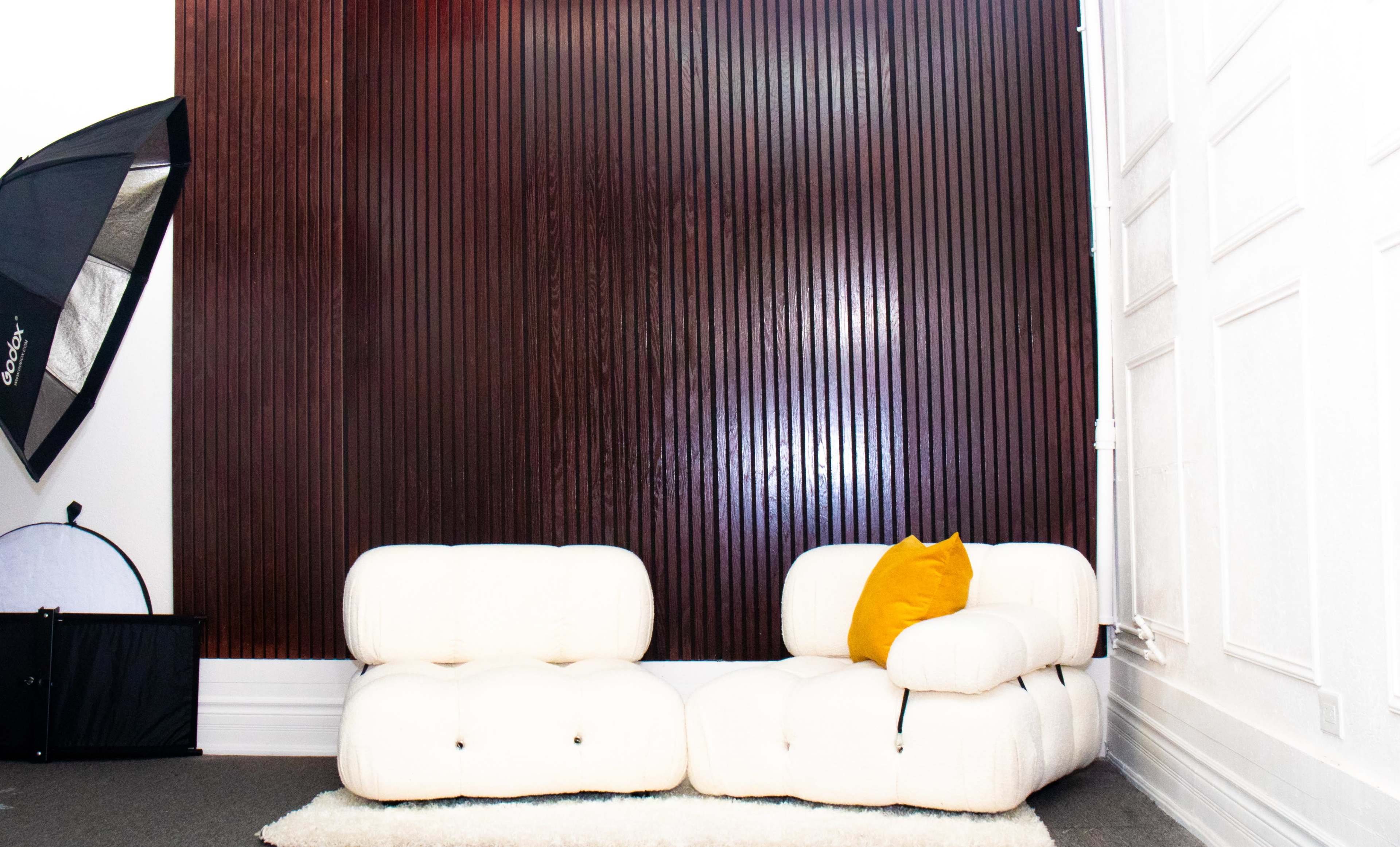 A modern, white, tufted sofa with a yellow cushion sits against a dark wooden slatted wall in a well-lit room.