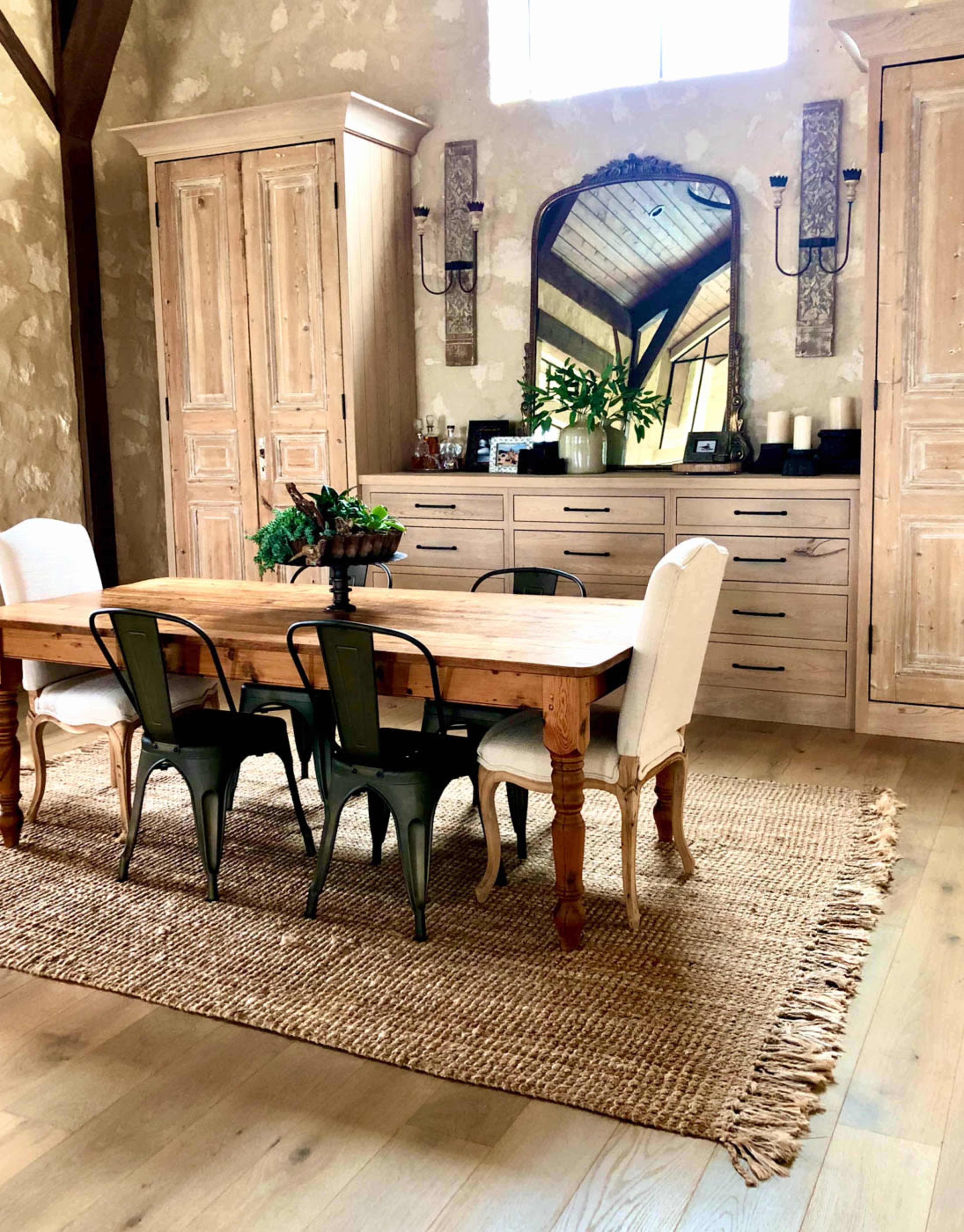 A rustic dining area features a wooden table surrounded by mixed seating, with a large mirror and storage cabinets against the wall.
