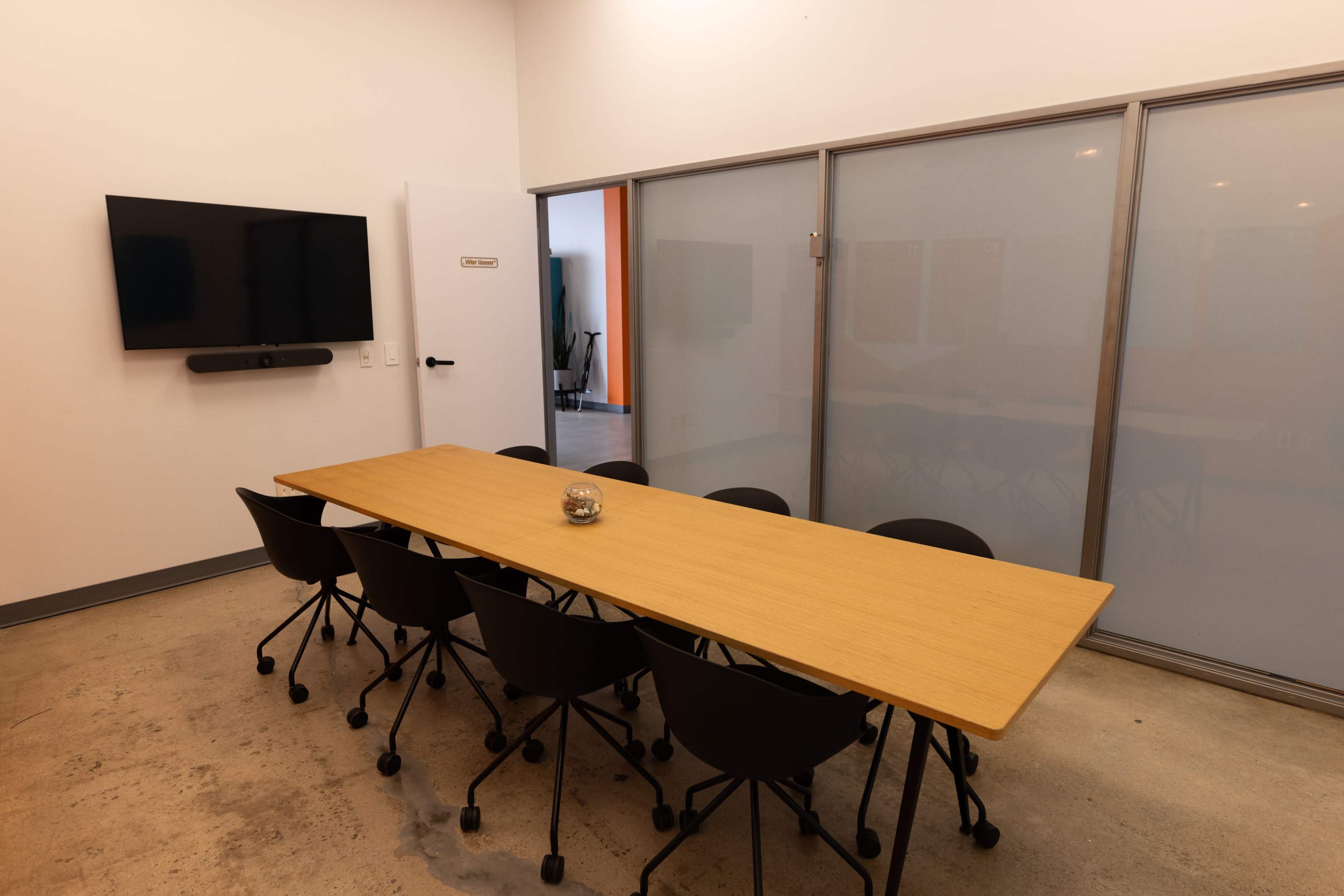 A rectangular wooden conference table with eight black rolling chairs is positioned in a meeting room with a television mounted on the wall and a glass partition.