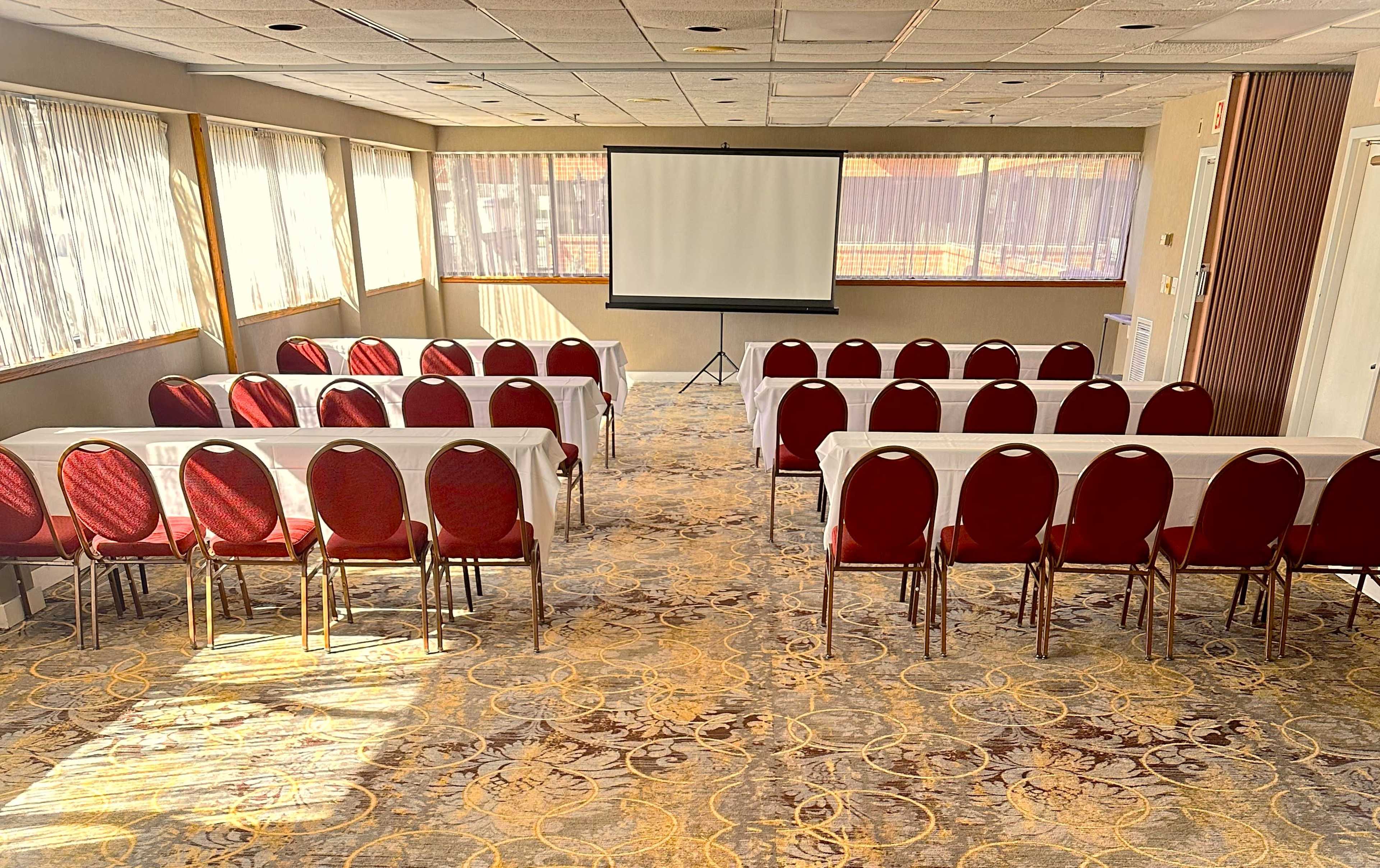 A meeting room is arranged with rows of red chairs and tables facing a projection screen.