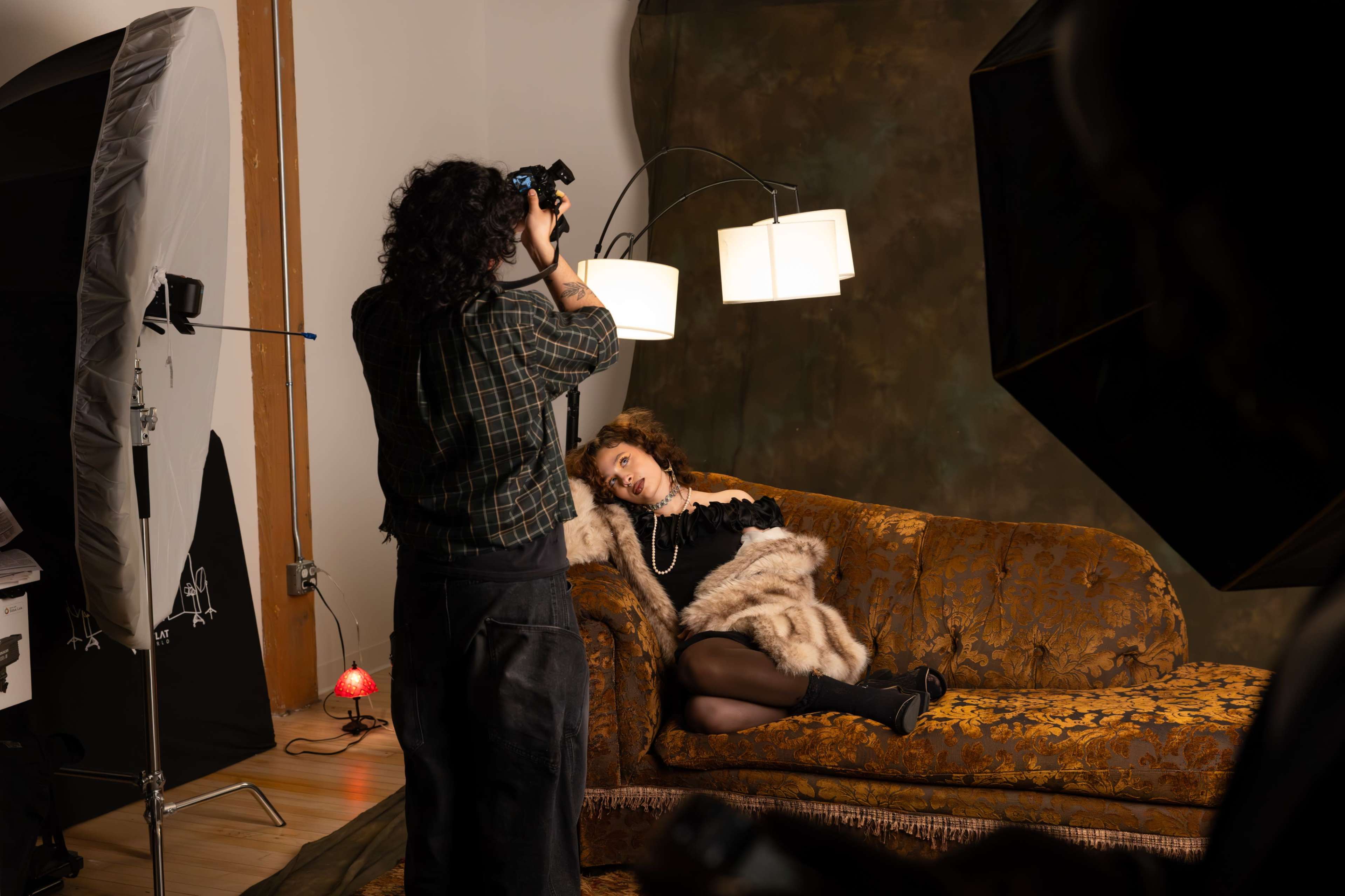 A woman poses on a vintage couch while a photographer captures her image in a studio setting.