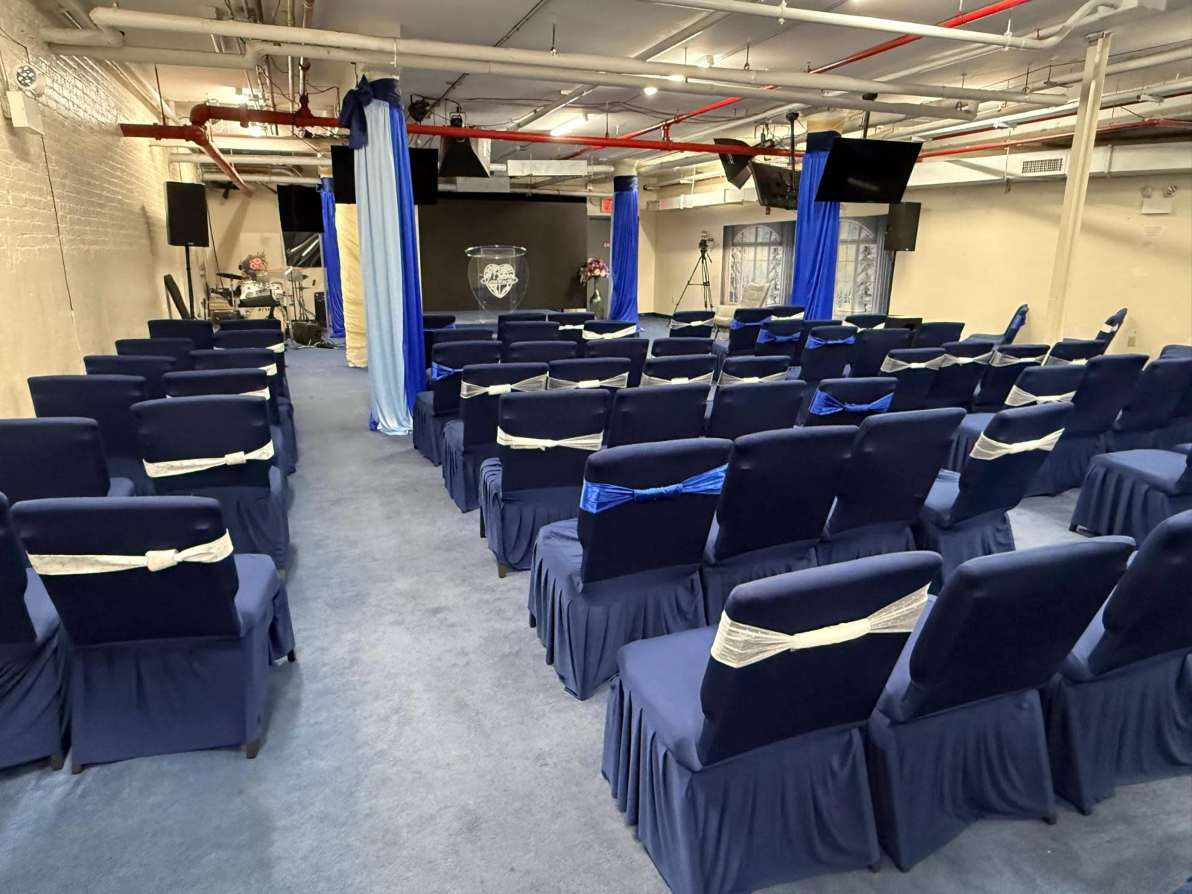 The image shows a conference room set up with rows of chairs covered in navy fabric and blue ribbons, facing a stage with equipment for a presentation in the background.