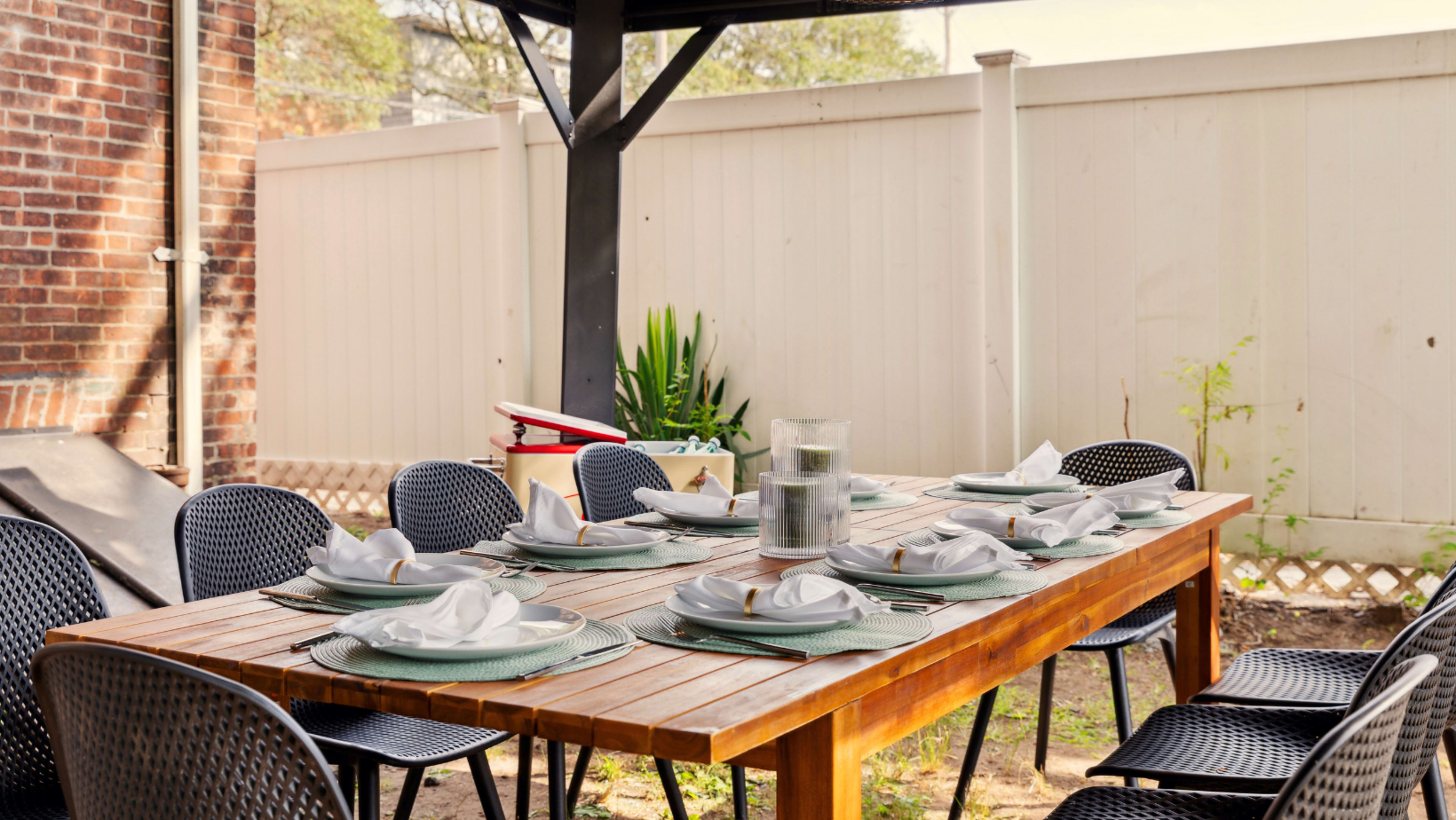 A wooden dining table is set outdoors with plates and folded napkins, surrounded by black chairs under a covered patio.