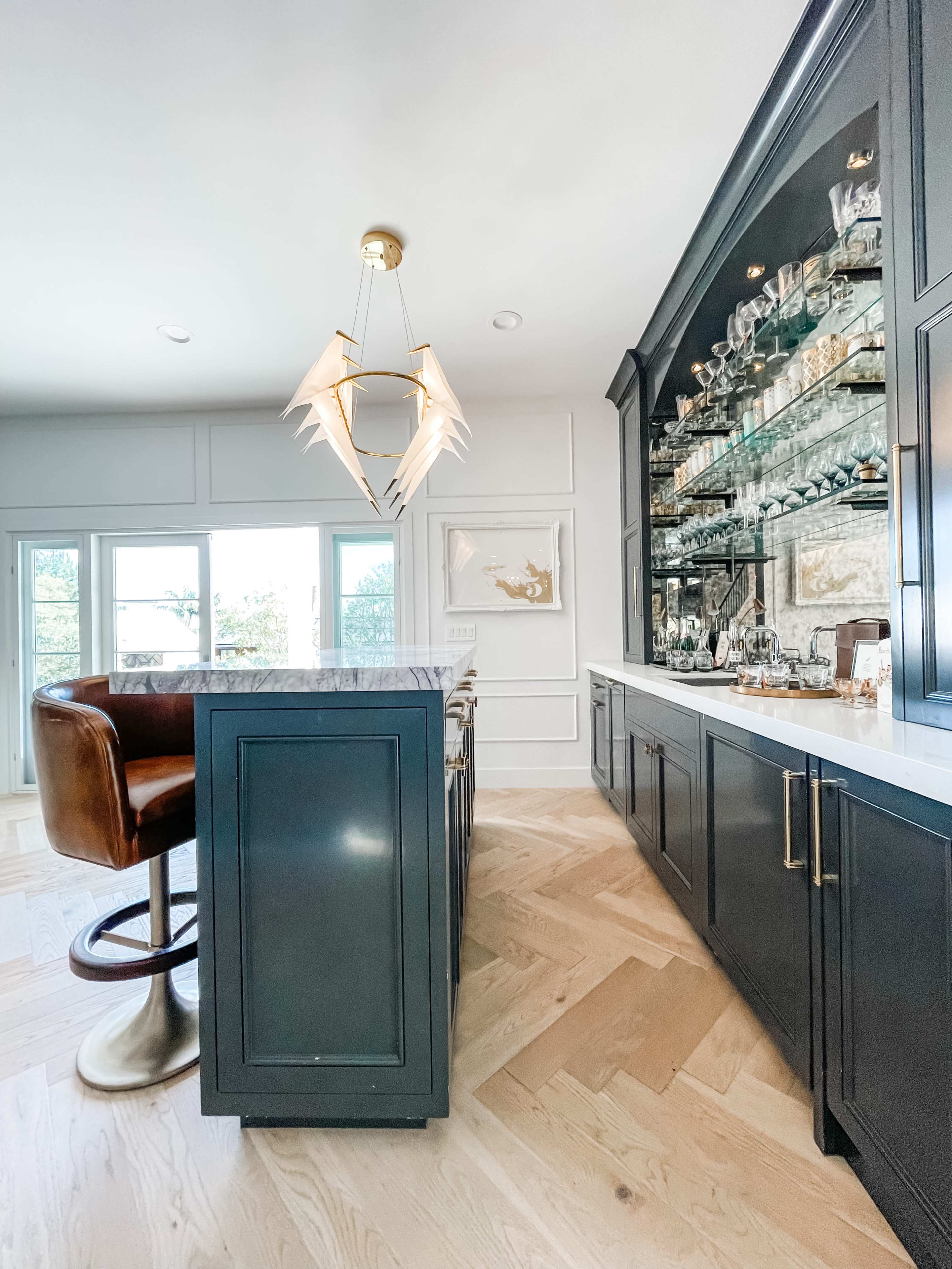 The image shows a modern kitchen with a dark cabinetry and a bar area featuring a high stool and a glass display cabinet filled with glassware.