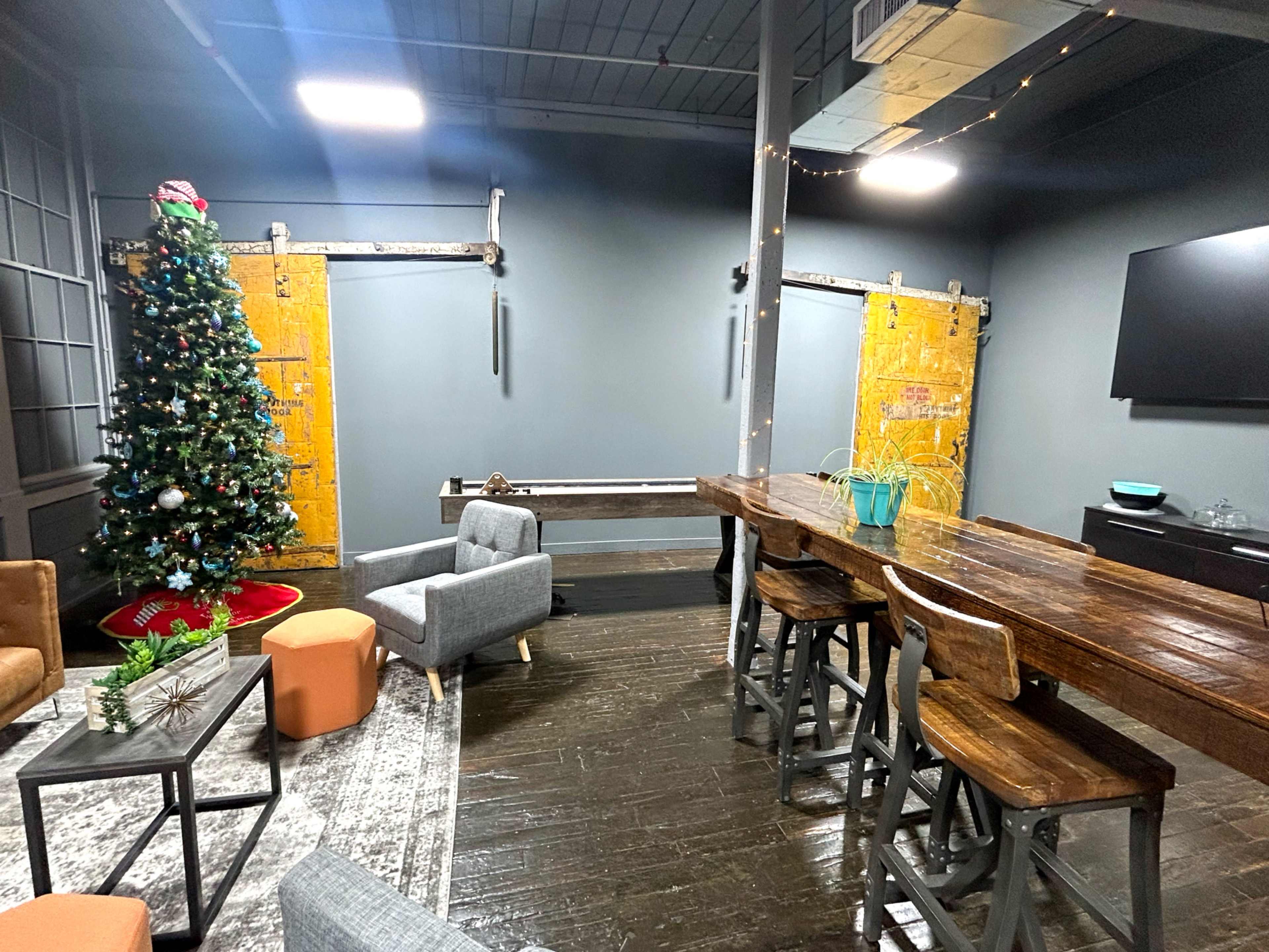The image shows a modern interior space featuring a large wooden table, gray seating, a decorated Christmas tree, and yellow sliding doors against a dark wall.