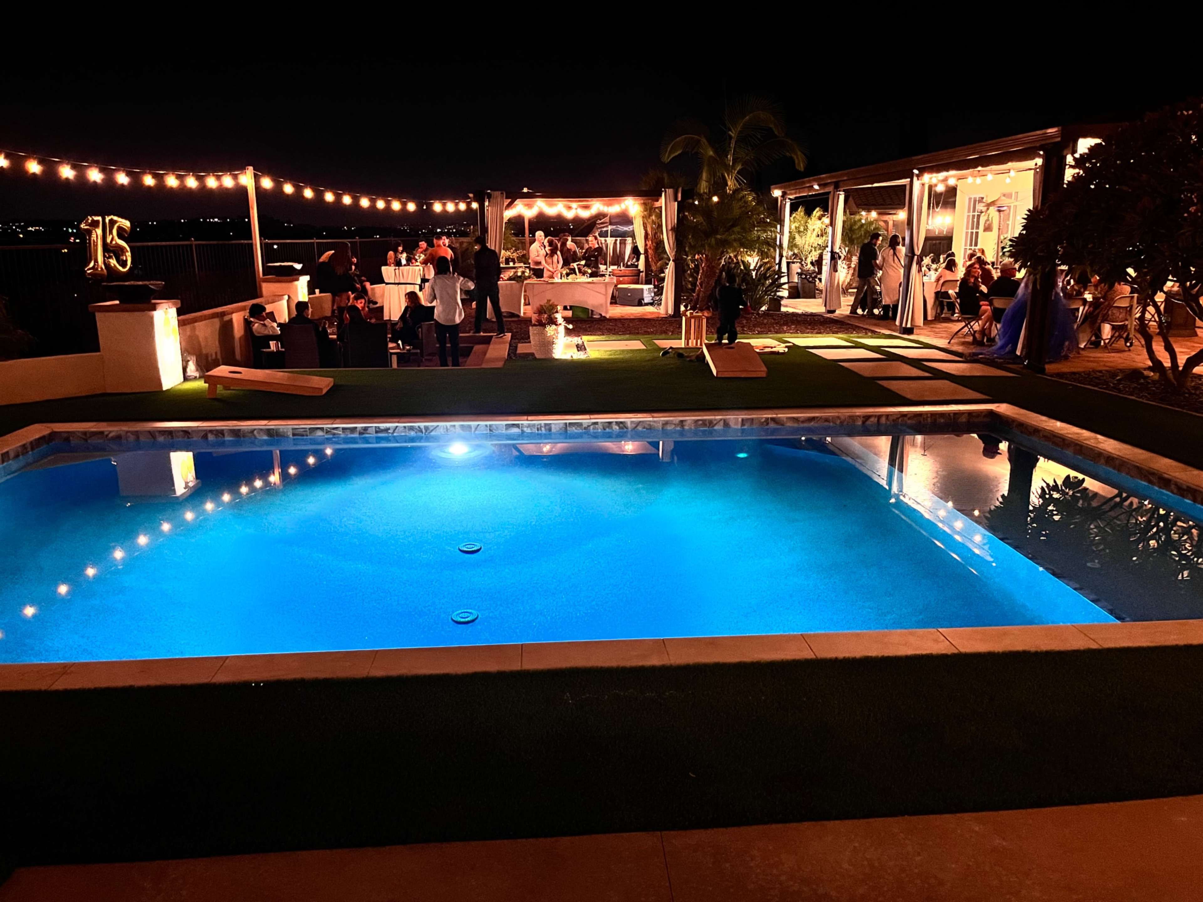 A brightly lit pool area is set for a nighttime celebration with string lights overhead and guests mingling by a decorated outdoor space.
