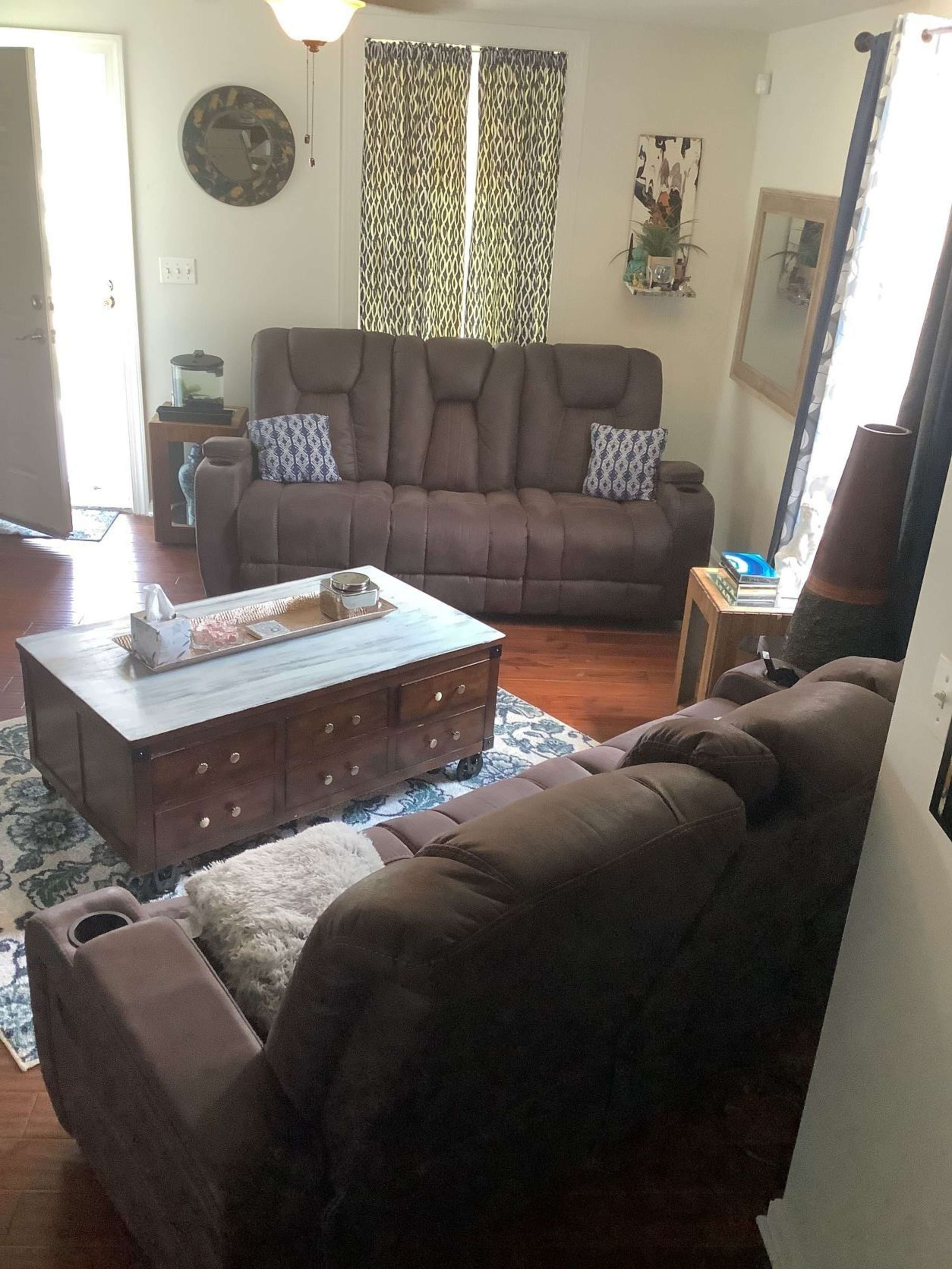 A living room with two brown couches, a wooden coffee table, and decor including curtains, a clock, and artwork on the walls.
