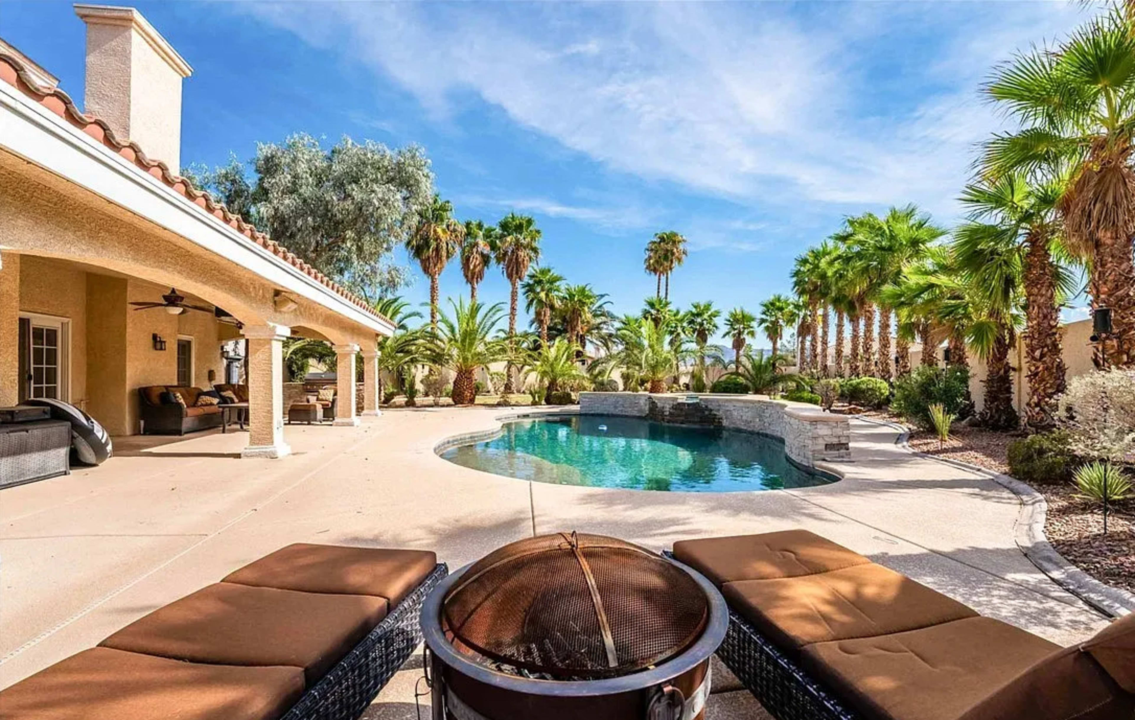 The image shows a landscaped backyard featuring a swimming pool surrounded by palm trees and outdoor seating areas.
