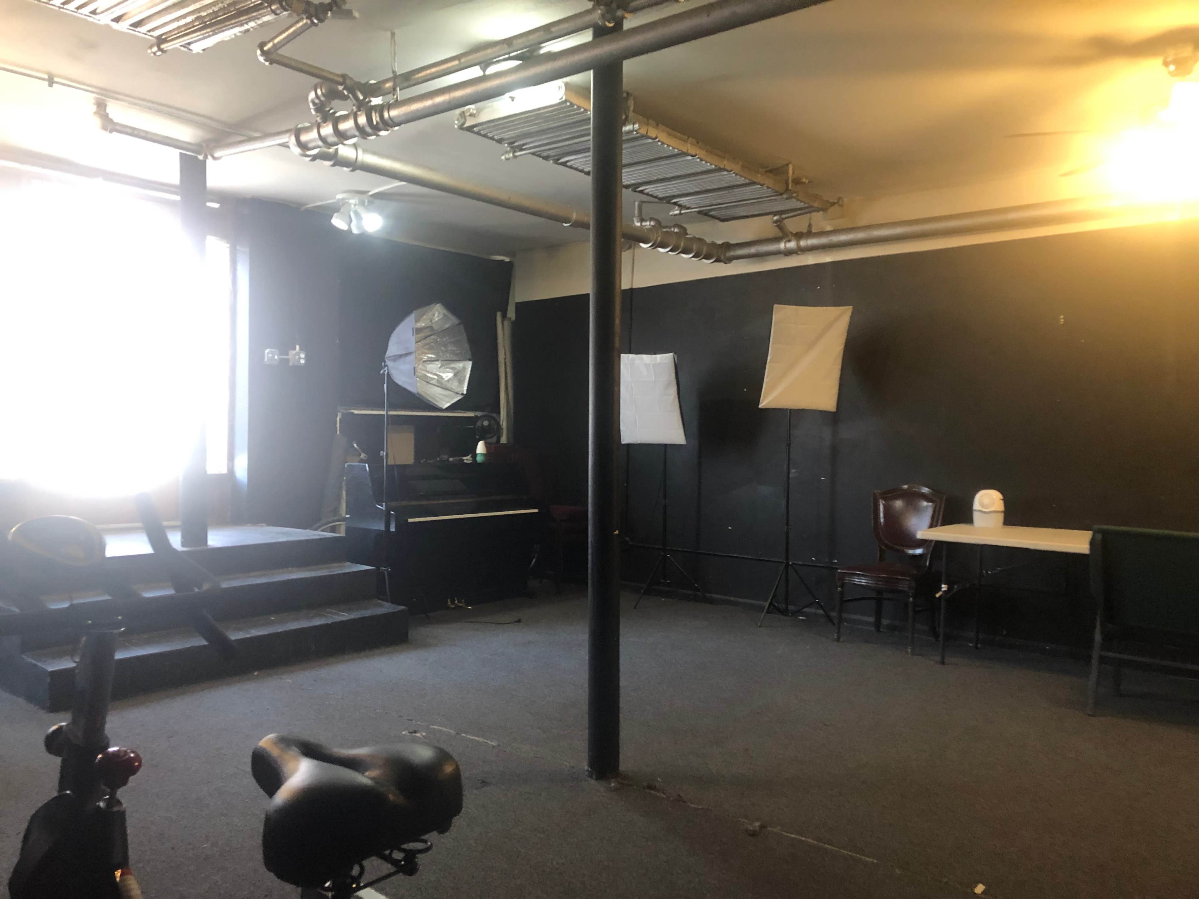 An empty room features a staircase, a piano, two light stands, a table with chairs, and a bicycle in the foreground.