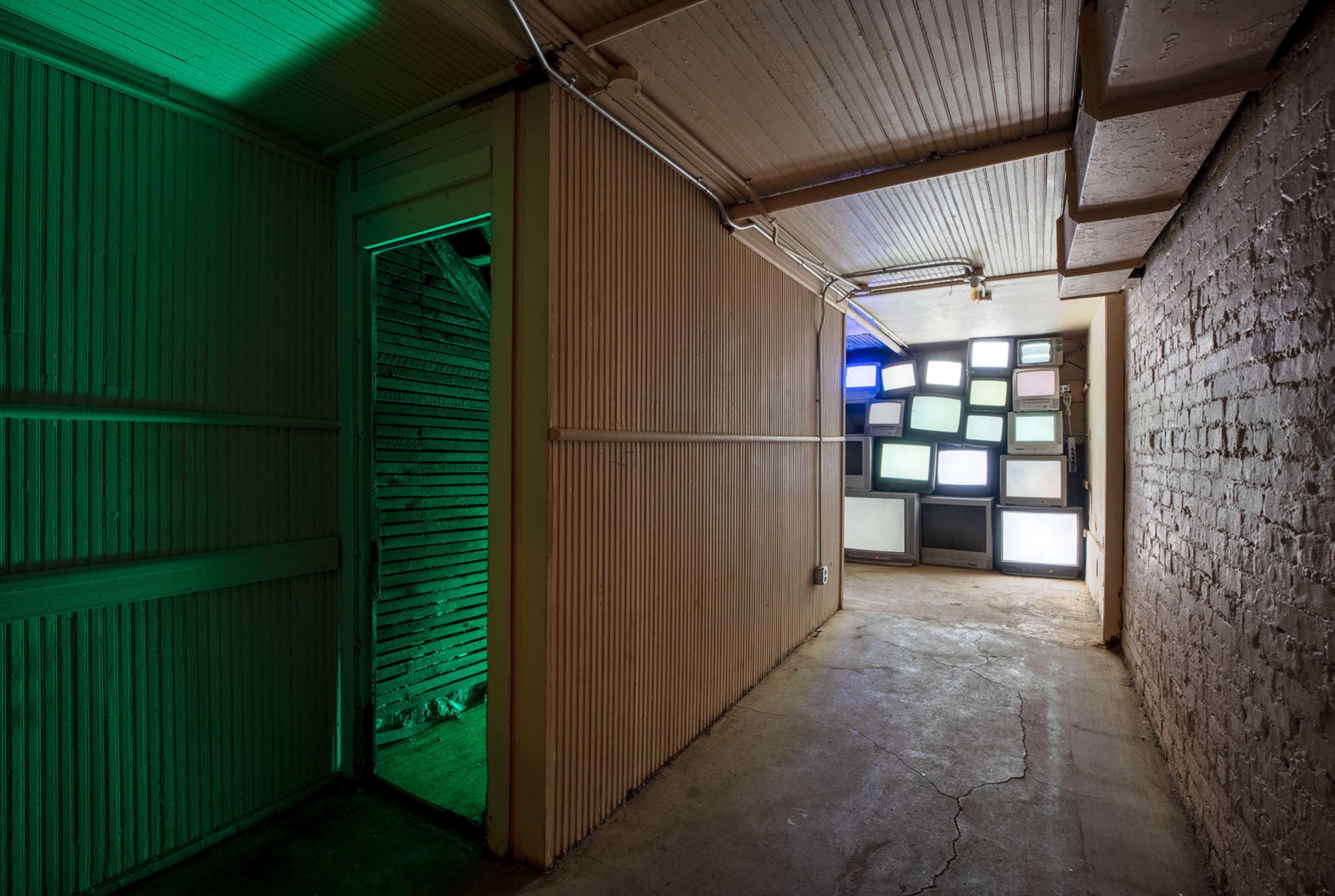 A narrow hallway leads to a wall of illuminated television screens, with a green-lit doorway on the left.