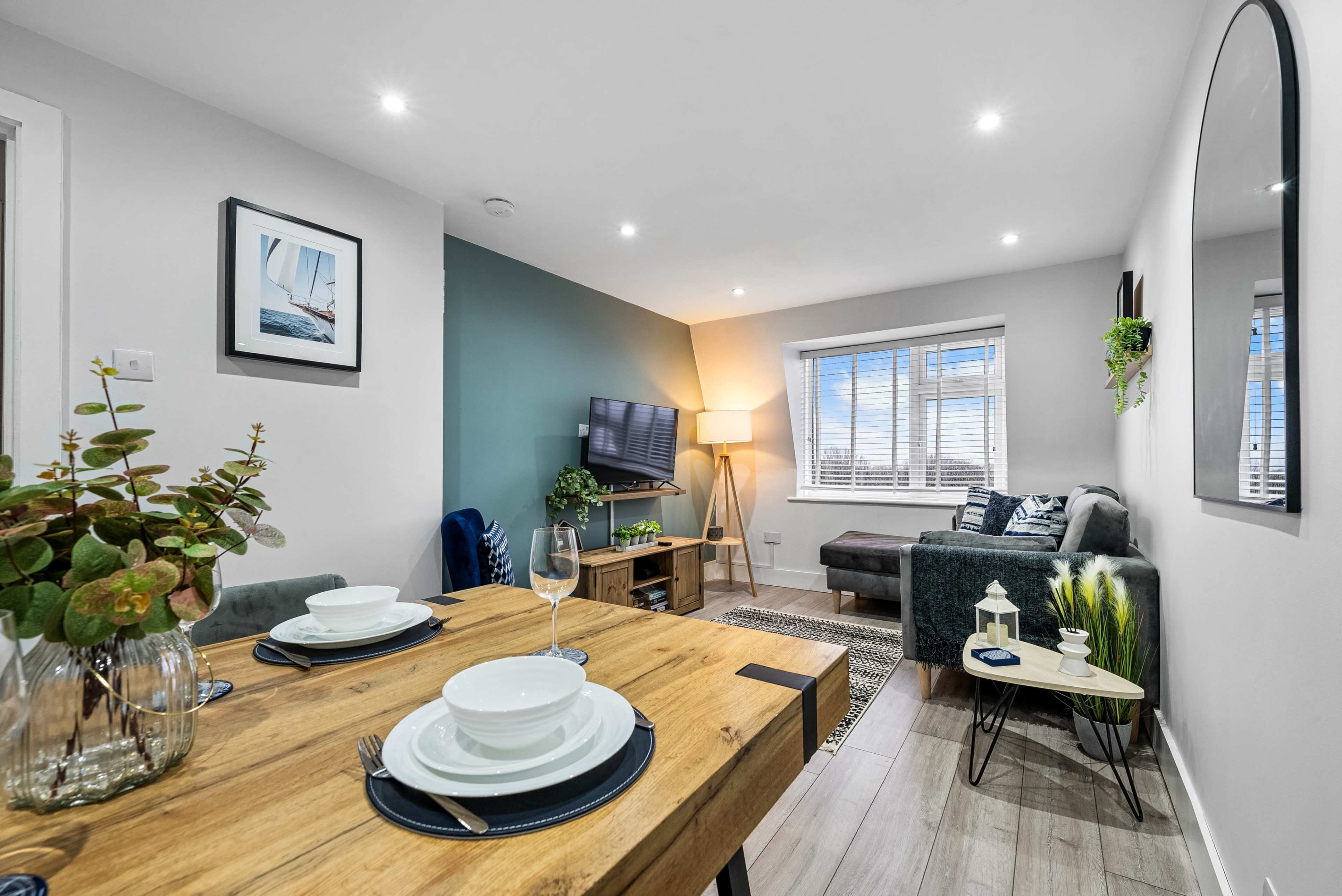 The image shows a modern living space with a dining area featuring a wooden table set for two, a cozy sofa in the corner, and a wall-mounted television.