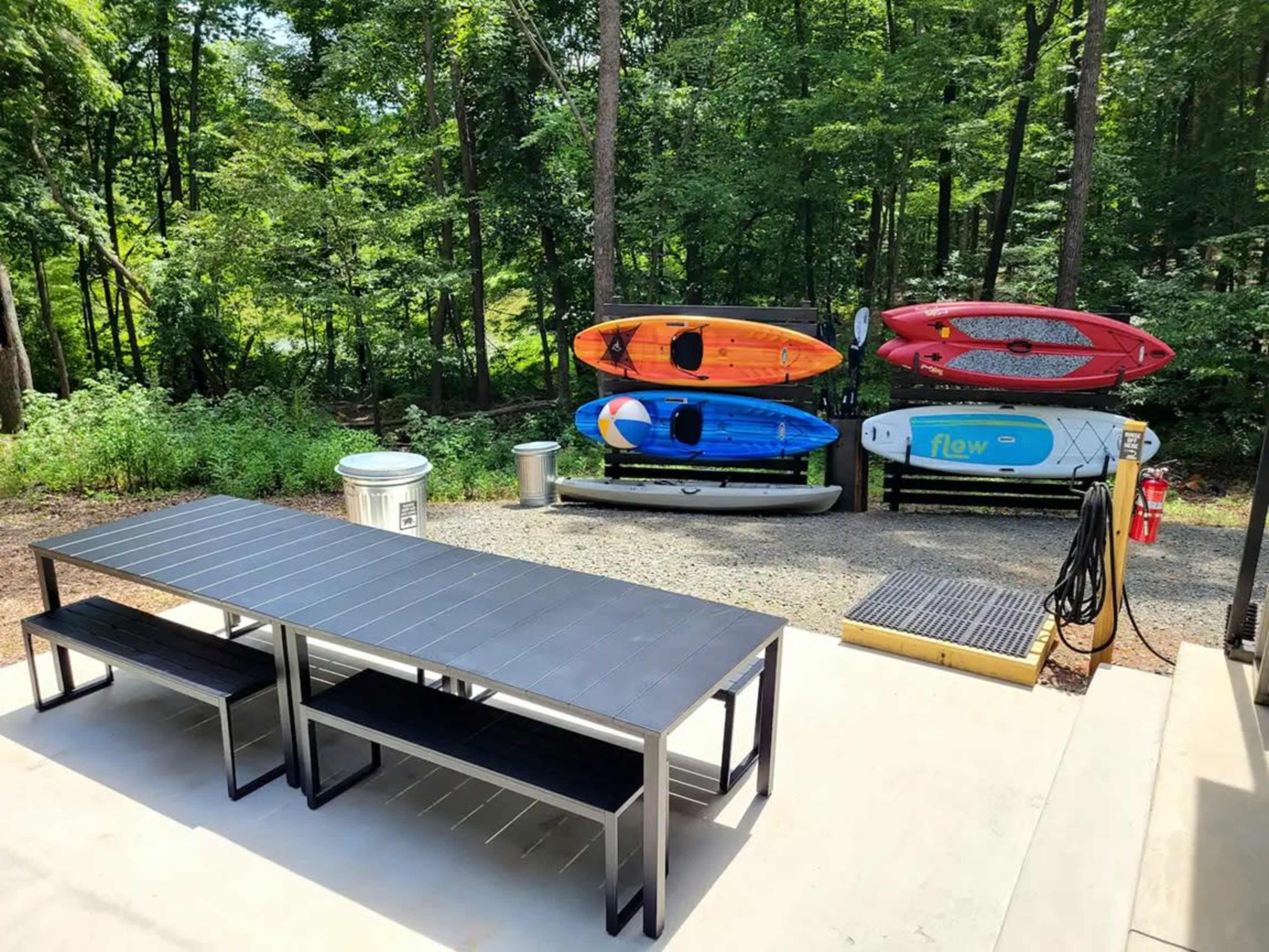 The image shows an outdoor patio area with a long table and benches, next to a display of colorful kayaks and a storage area.