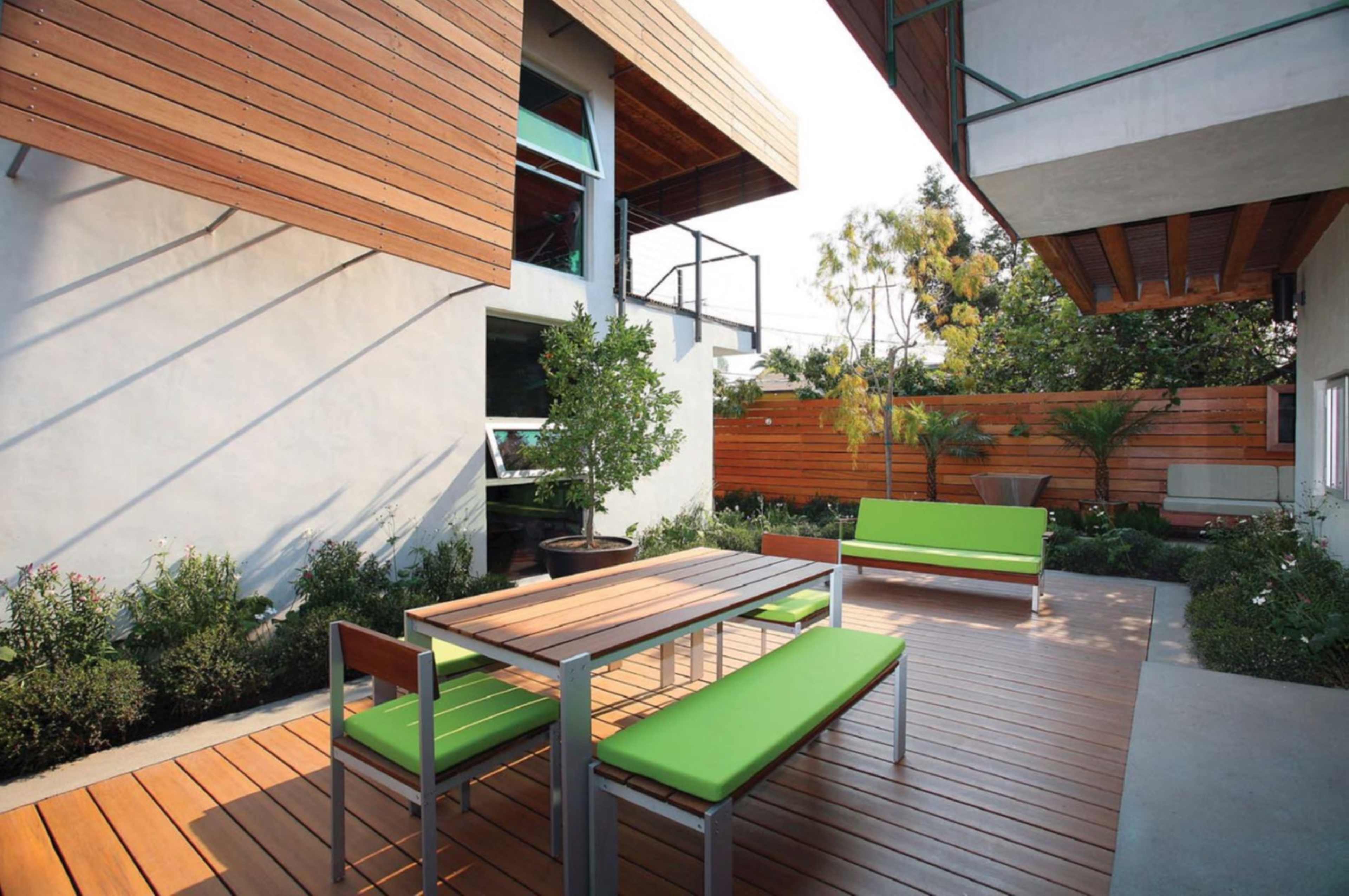 The image shows a modern outdoor patio with a wooden table and green benches surrounded by landscaped plants and wooden fencing.