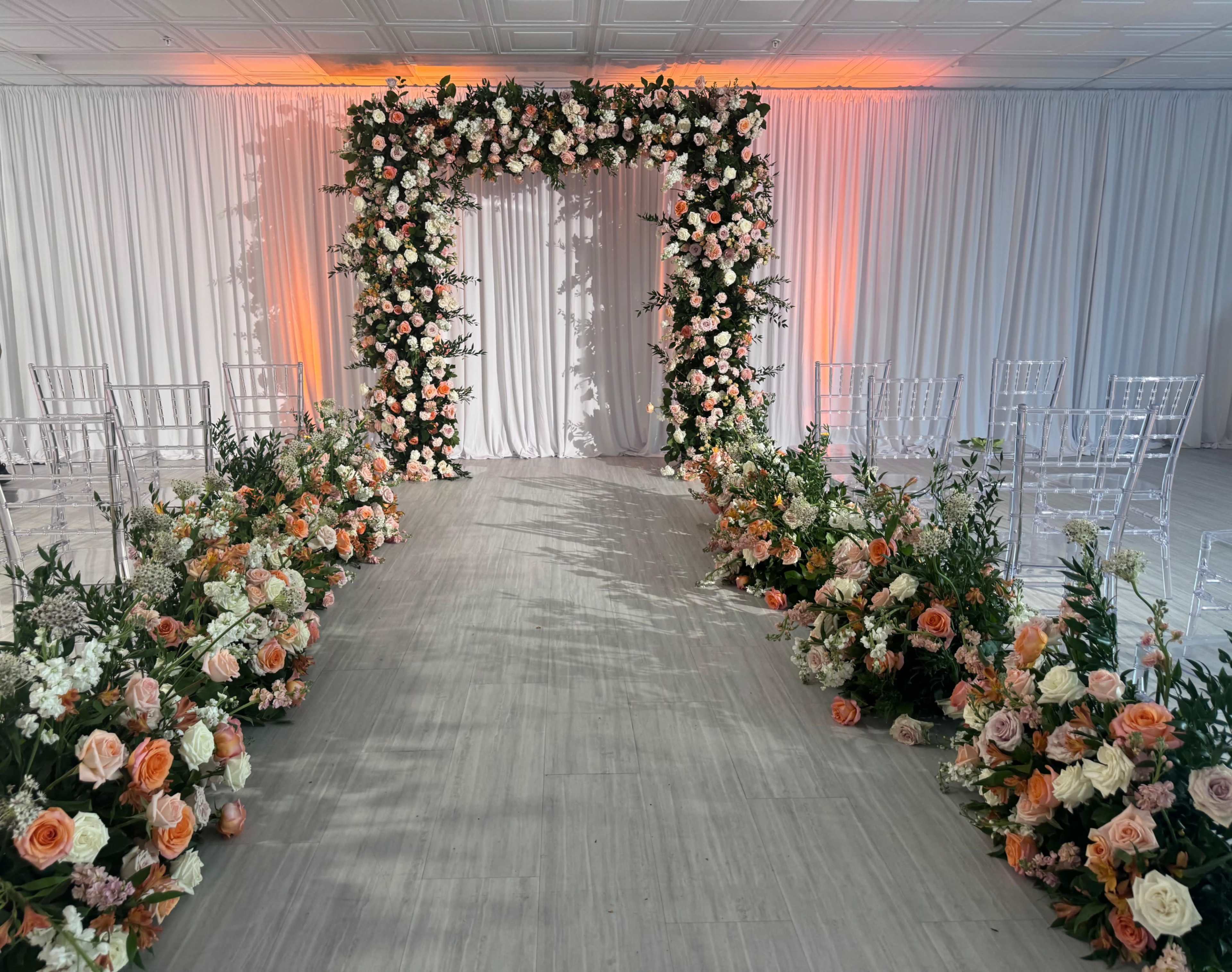 A wedding ceremony setup featuring a floral archway adorned with roses and greenery, flanked by rows of clear chairs and floral arrangements lining a central aisle.