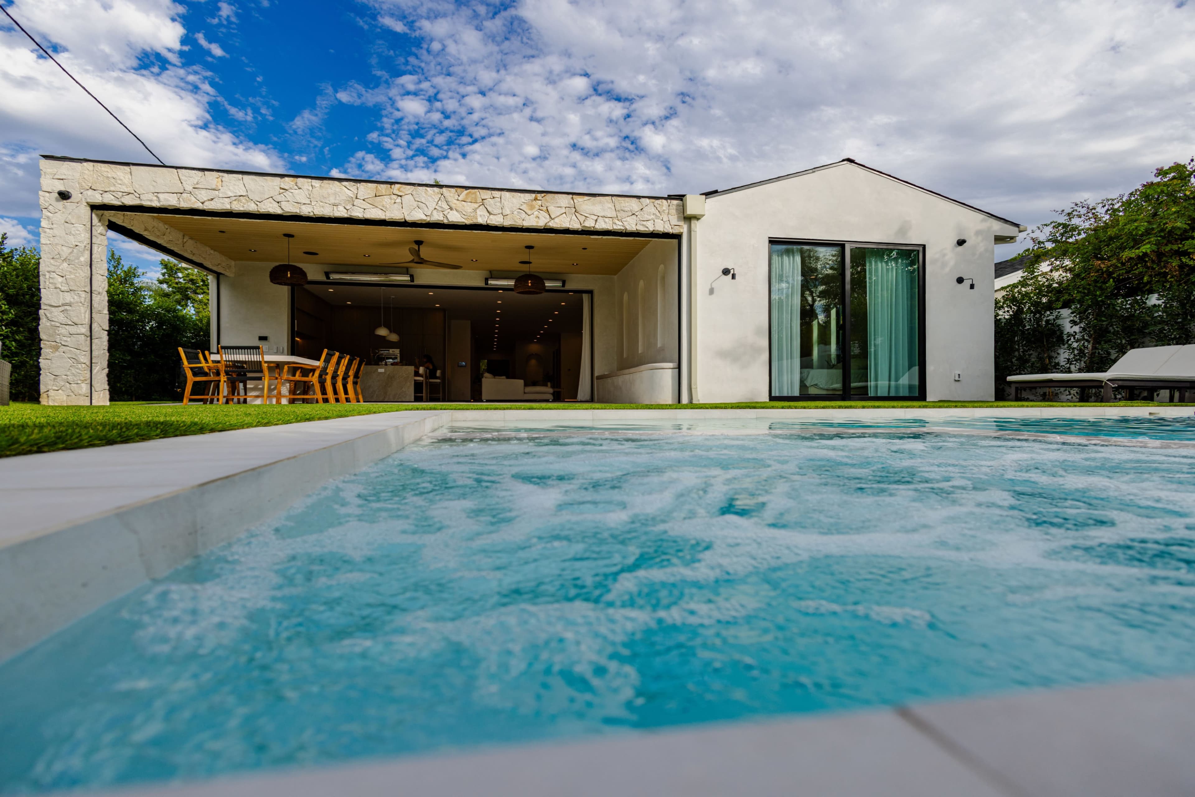 A modern house with large glass doors overlooks a swimming pool surrounded by grass and a stone patio under a partly cloudy sky.