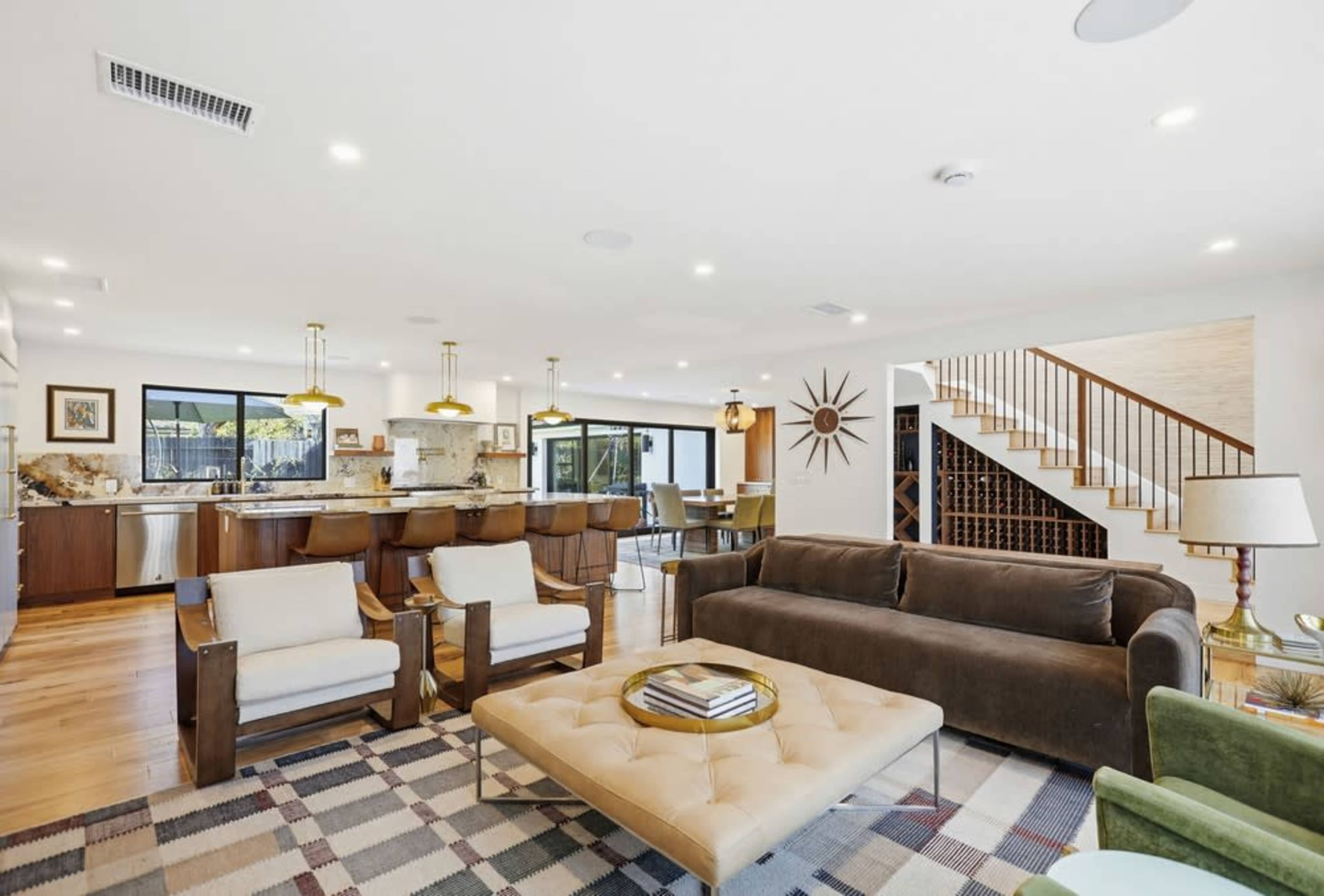 The image shows a modern open-concept living space featuring a sitting area with a brown sofa, two armchairs, a coffee table, and a kitchen area with bar seating.