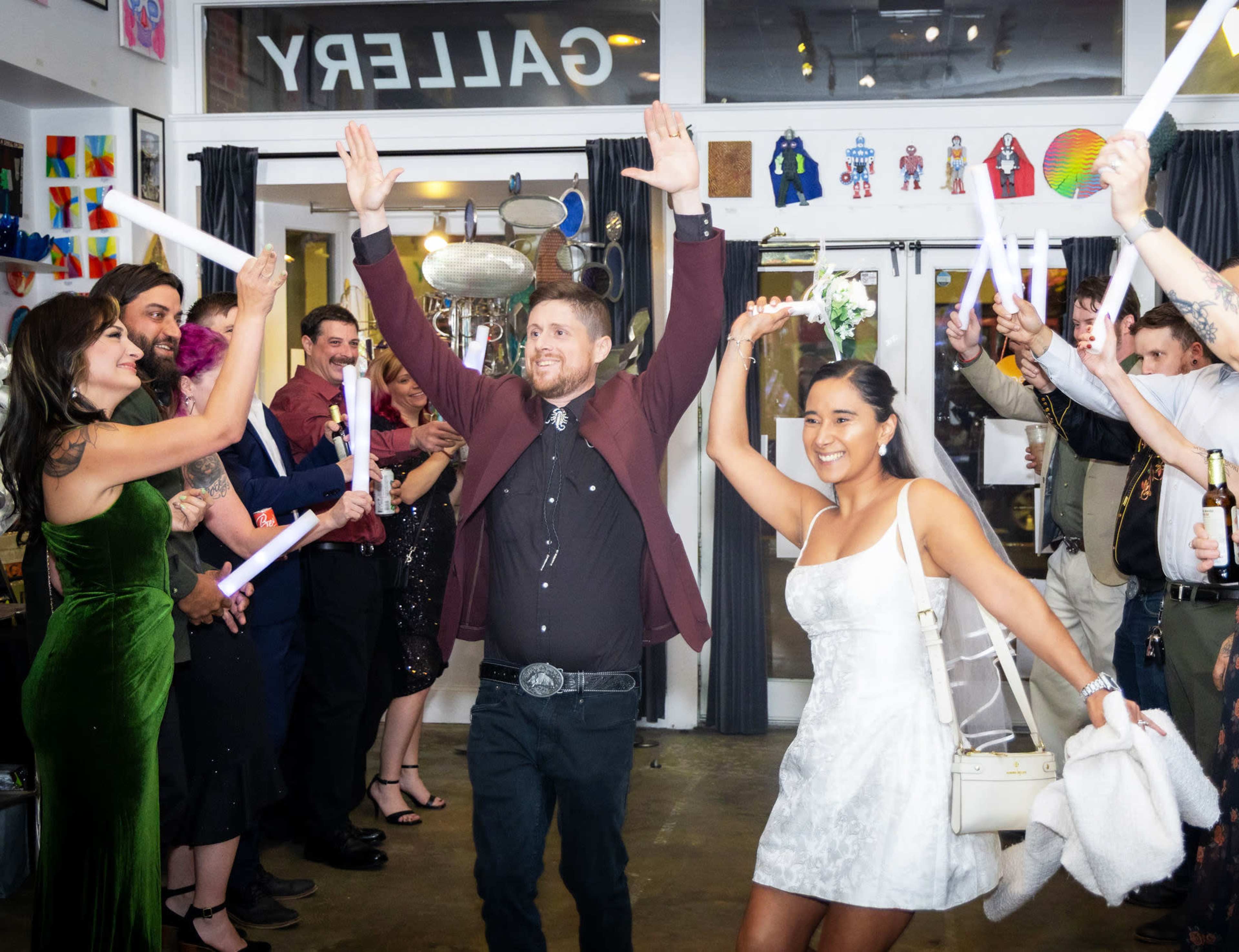 A group of people celebrate with raised arms and glowing sticks in an art gallery, while a couple leads the festivities at the entrance.