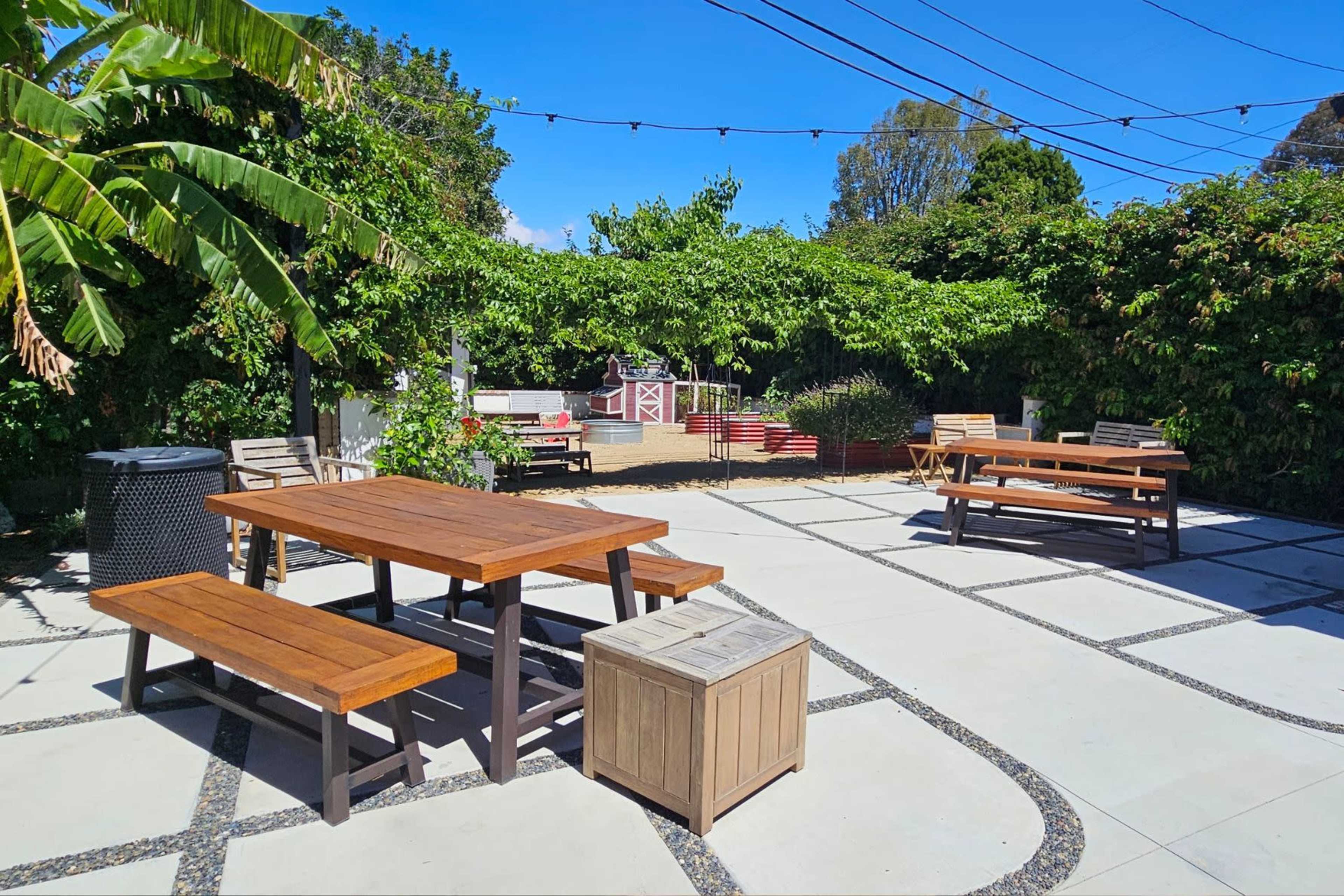 The image shows a well-designed outdoor patio area with wooden tables and benches surrounded by greenery and string lights.