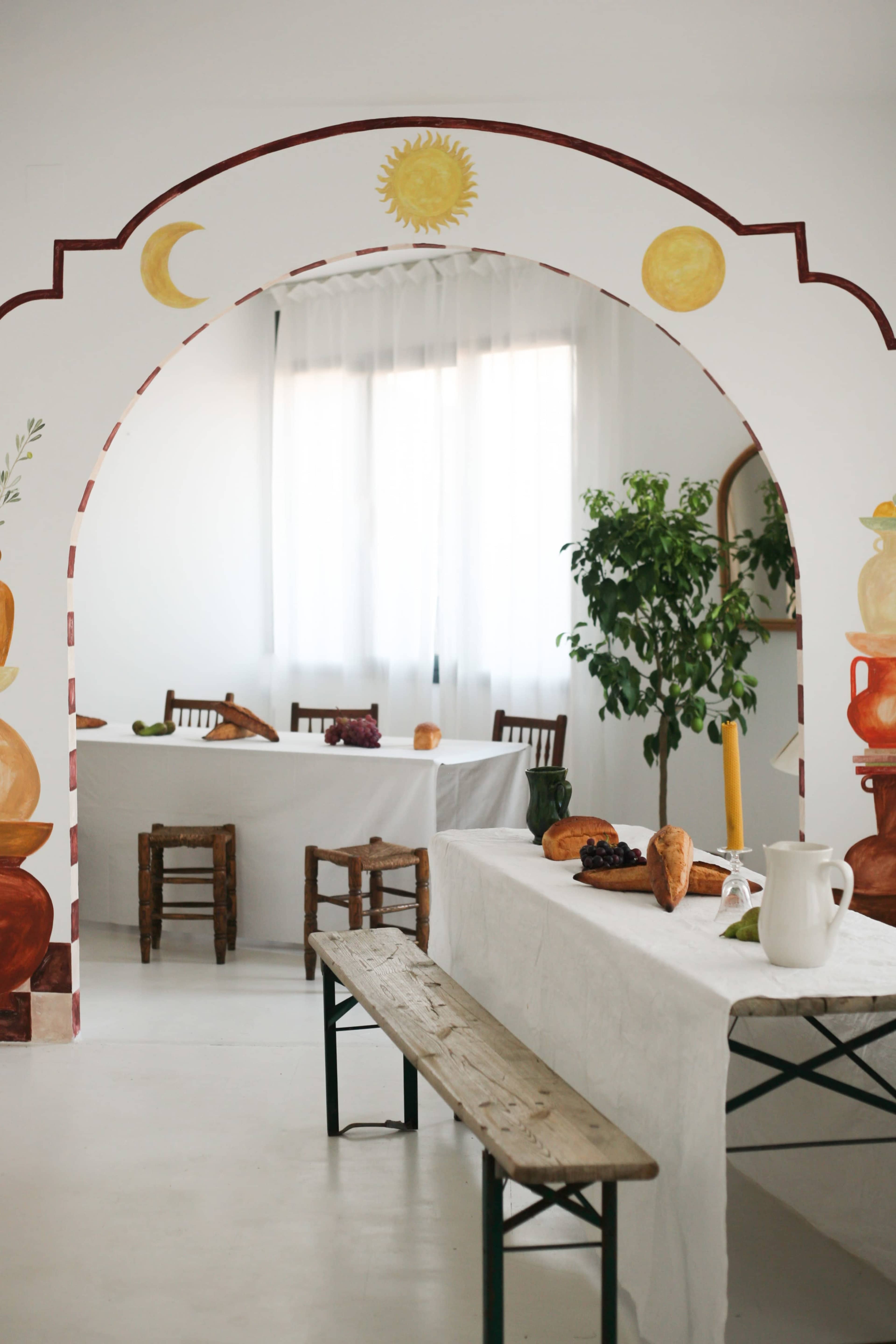 A bright, minimalistic dining space features a large archway with painted celestial designs, leading to tables adorned with white tablecloths and simple decor.