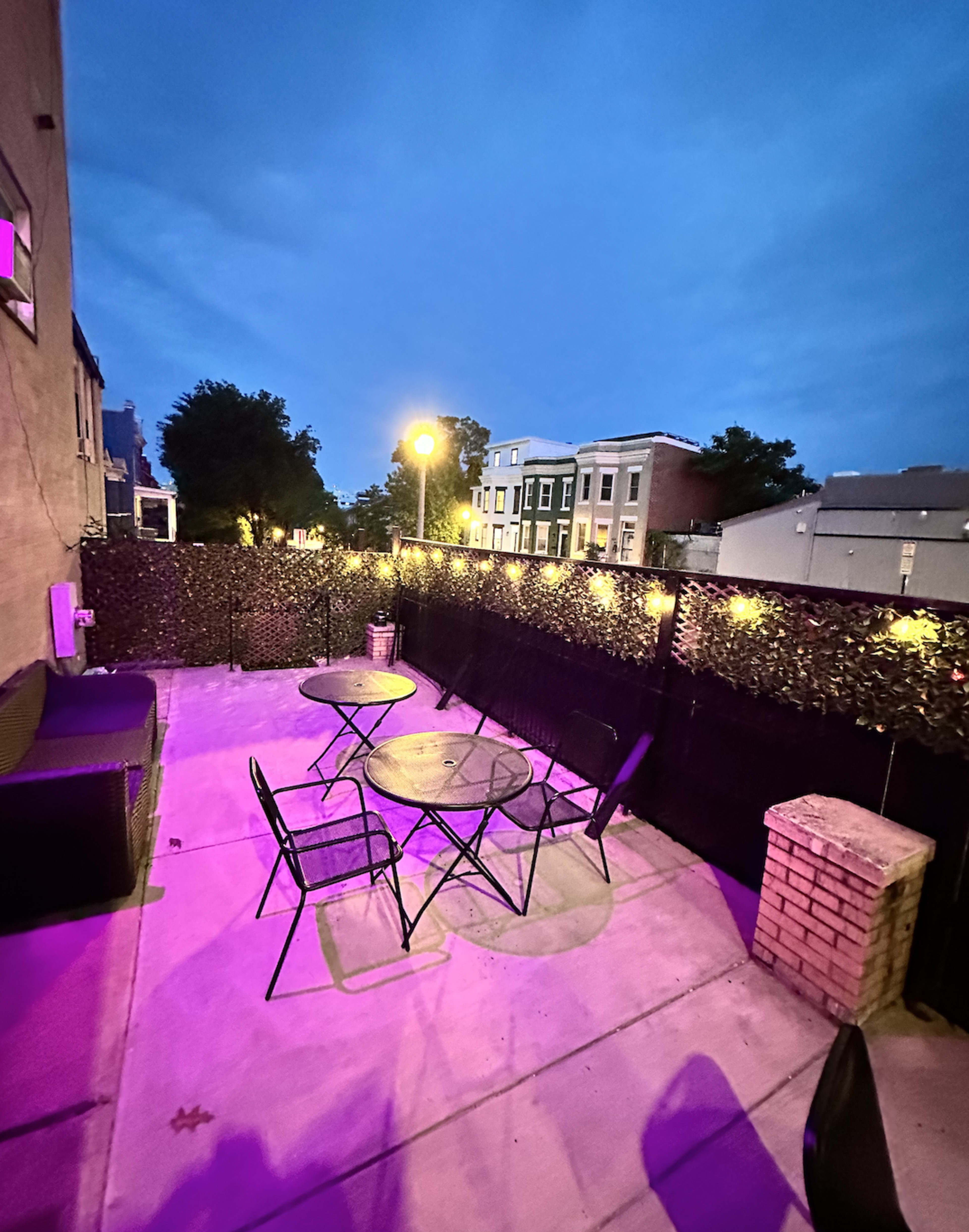 The image shows a patio area with two circular tables and four chairs, surrounded by a privacy wall and illuminated by purple lighting at dusk.