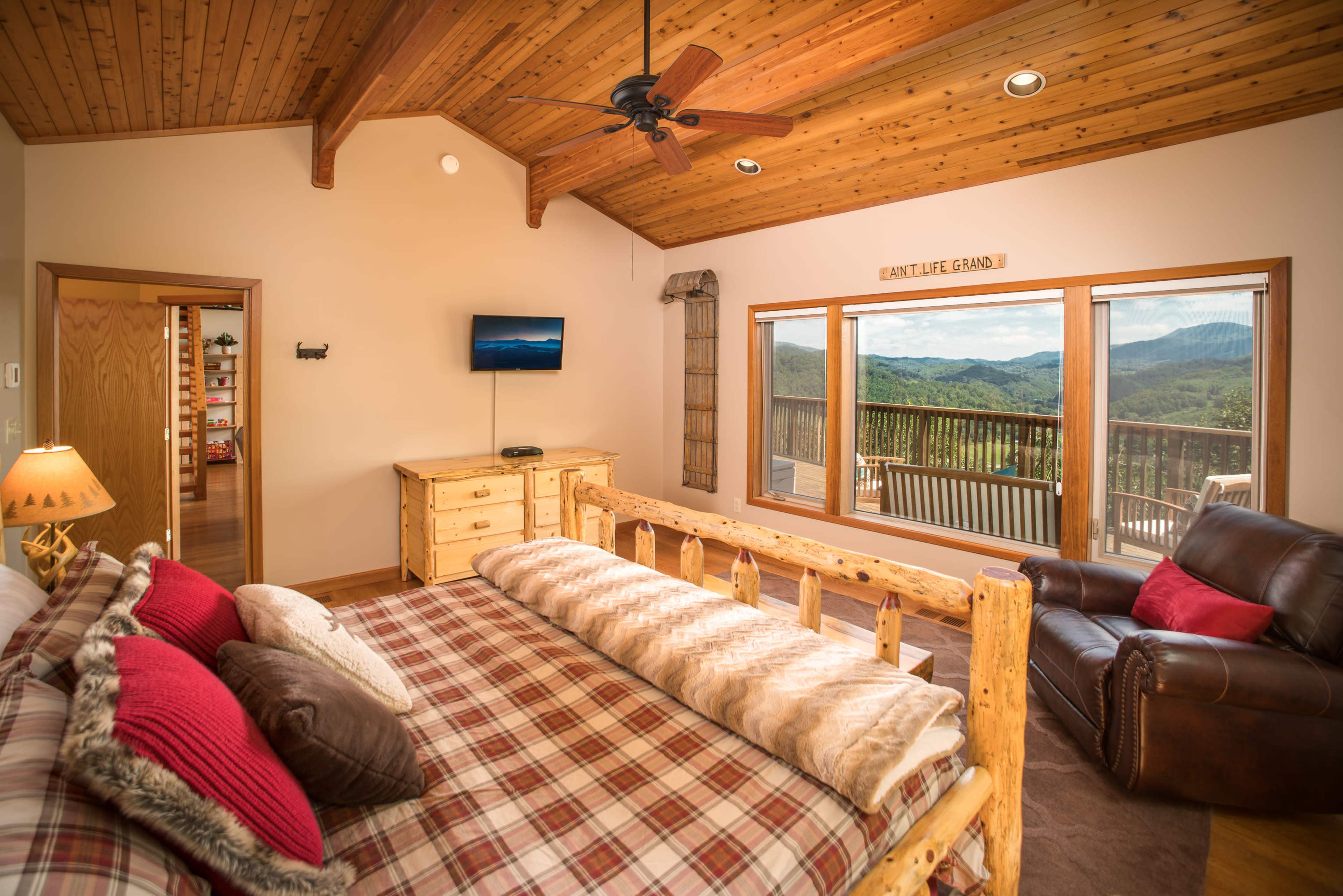A cozy bedroom with a wooden ceiling, a log bed covered with a plaid blanket, a dark leather chair, and large windows revealing a mountain view.