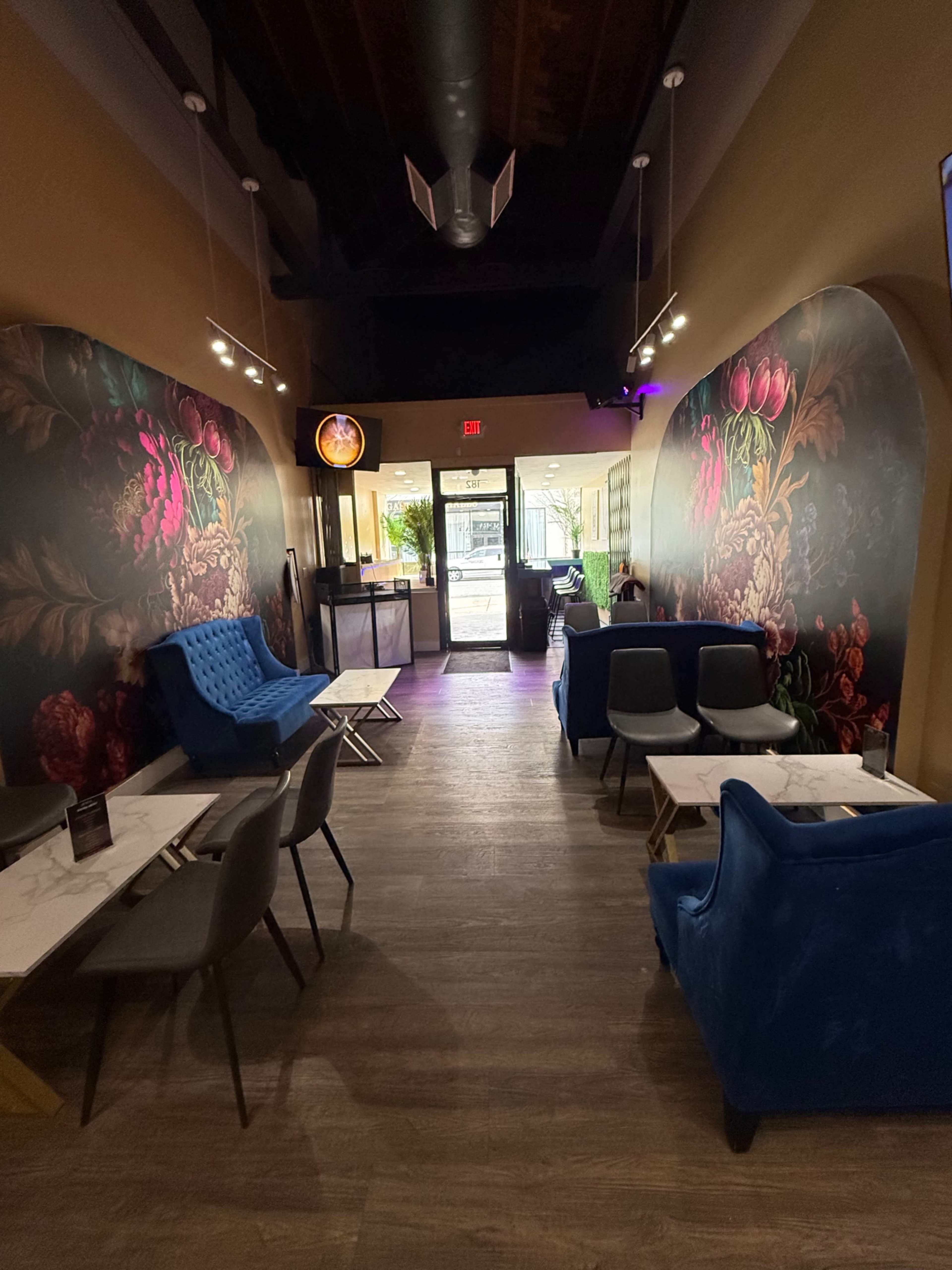 The image shows a stylish café interior featuring floral wallpaper, blue chairs, and tables arranged along the sides of a long corridor leading to an entrance.