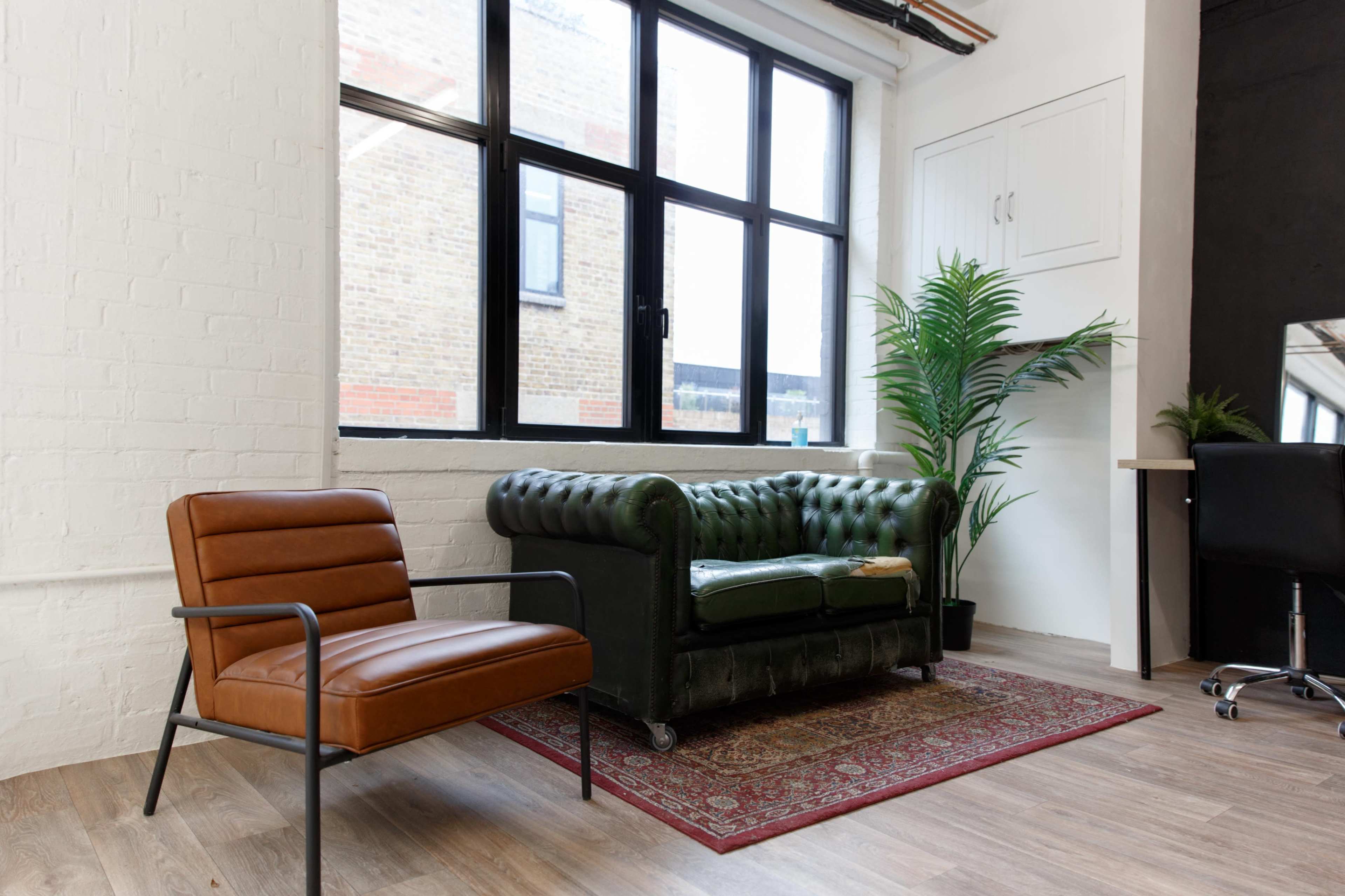 A modern room featuring a green leather chesterfield sofa, a brown armchair, large windows, and a potted plant.
