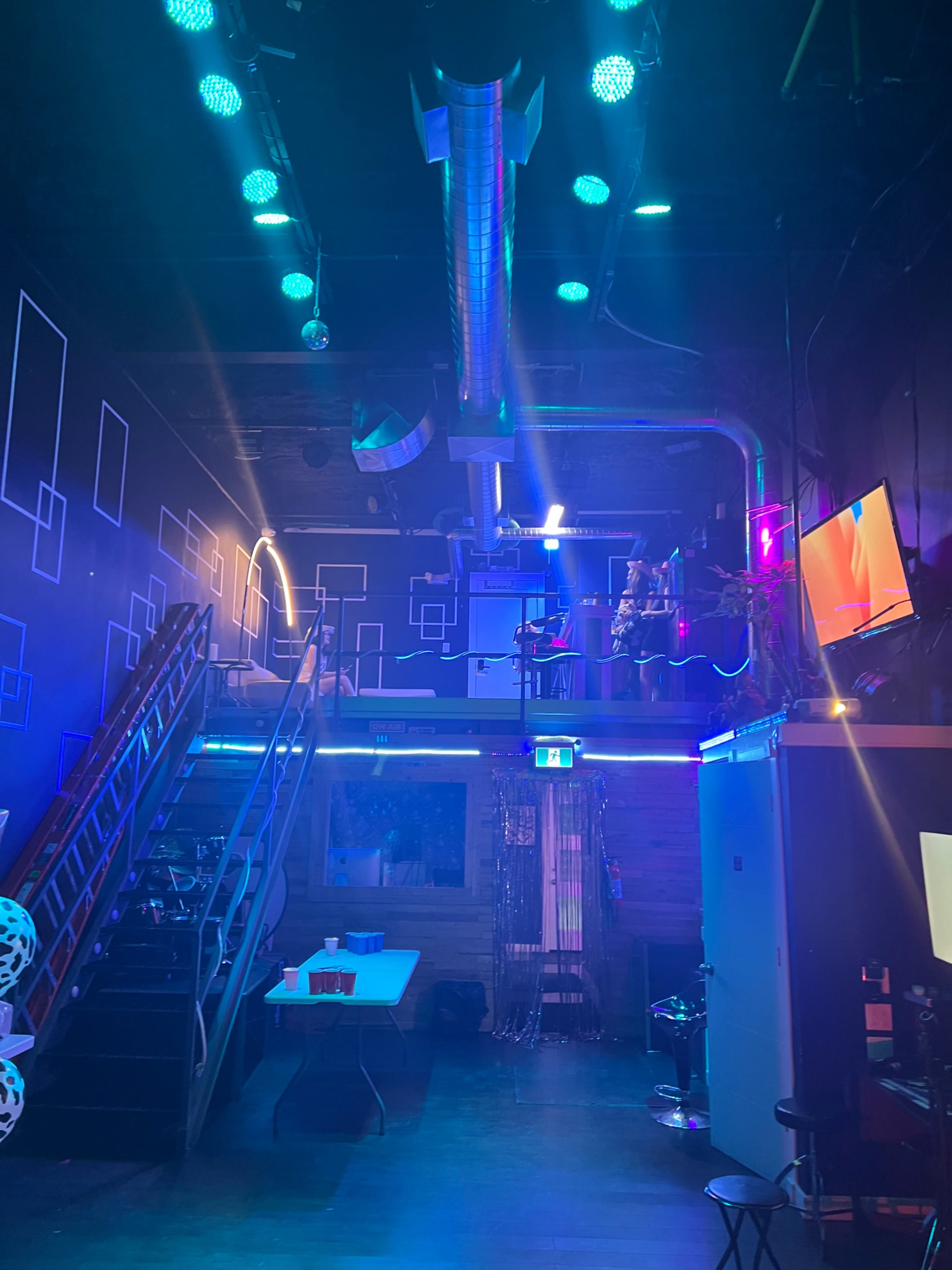 The interior of a dimly lit entertainment space features colorful lighting, a staircase leading to an upper level, and various equipment scattered around.
