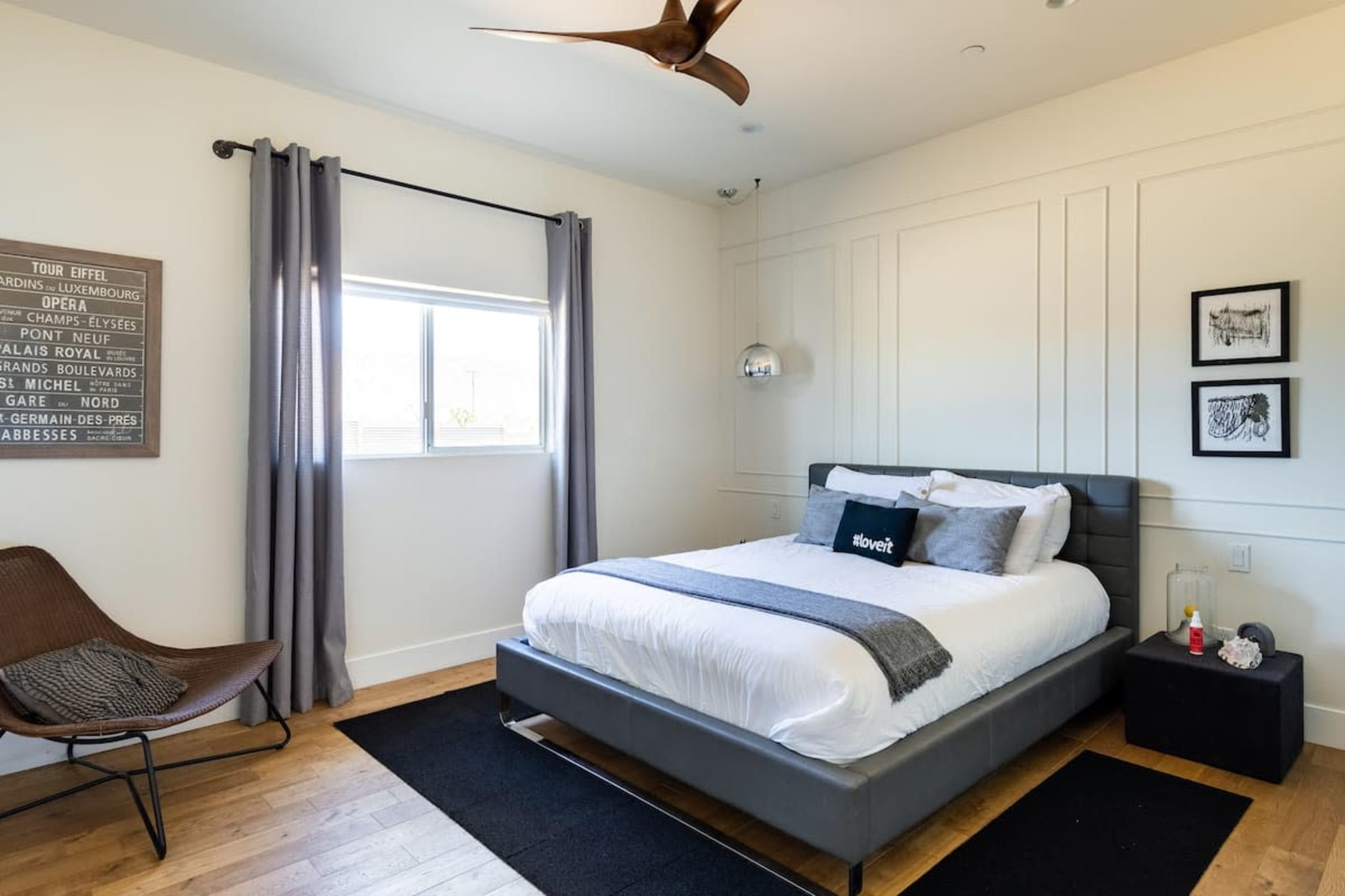 A modern bedroom features a gray upholstered bed with white linens, a wooden floor, a large window with curtains, and minimalist decor on the walls.