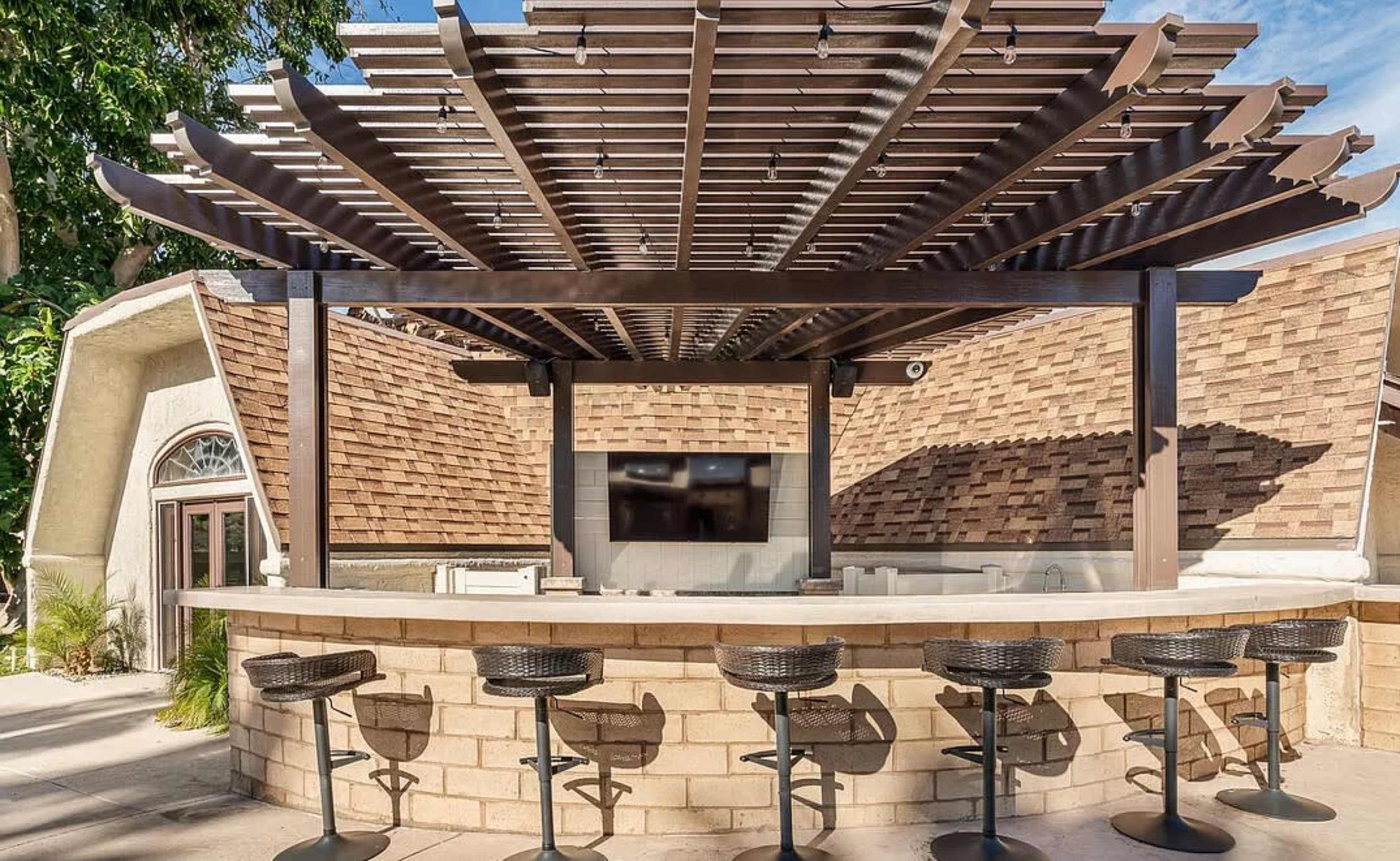 The image shows an outdoor bar with a wooden pergola, featuring a circular counter and several black wicker stools.