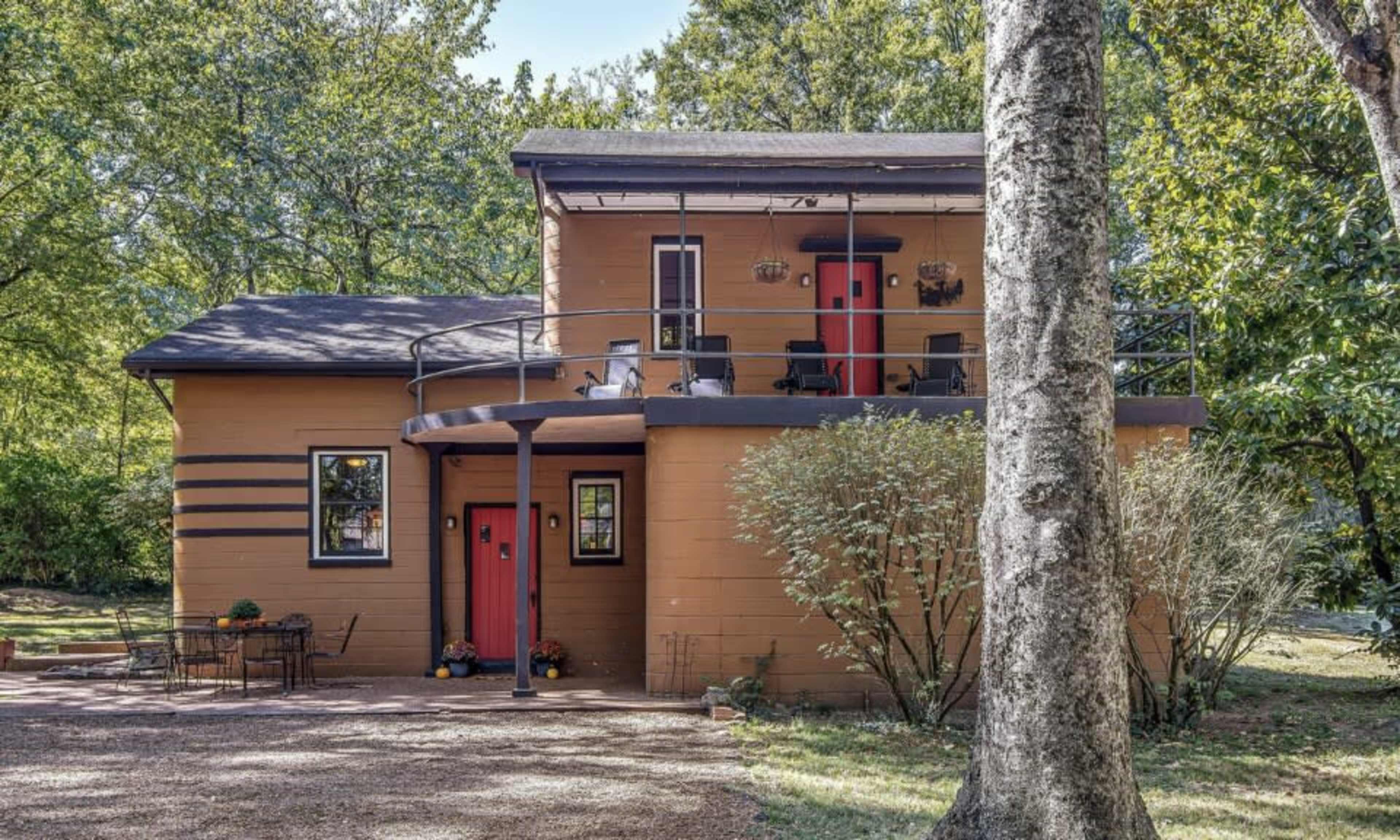 A two-story house with a brown exterior and red doors stands amidst trees, featuring a balcony and a gravel walkway.