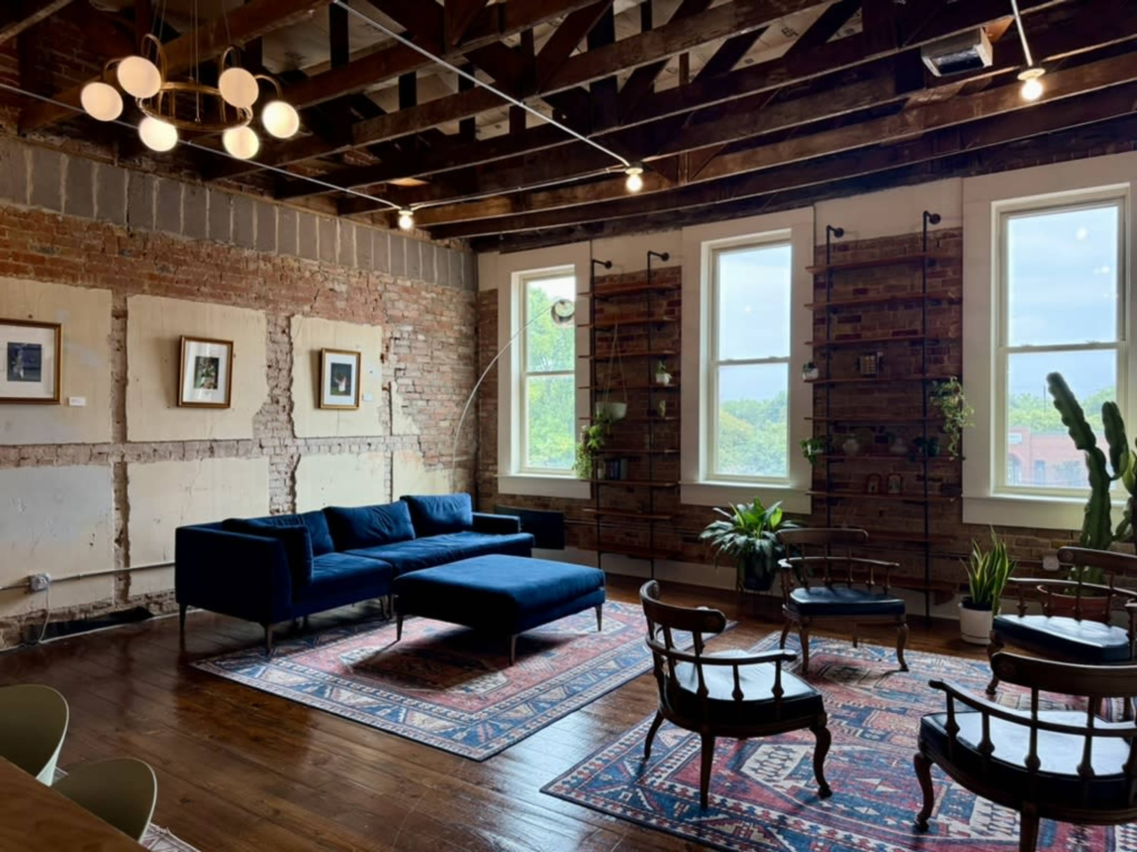 The image shows a spacious loft-style living room with exposed brick walls, large windows, and a combination of modern and vintage furniture, including a blue sectional sofa and wooden chairs.
