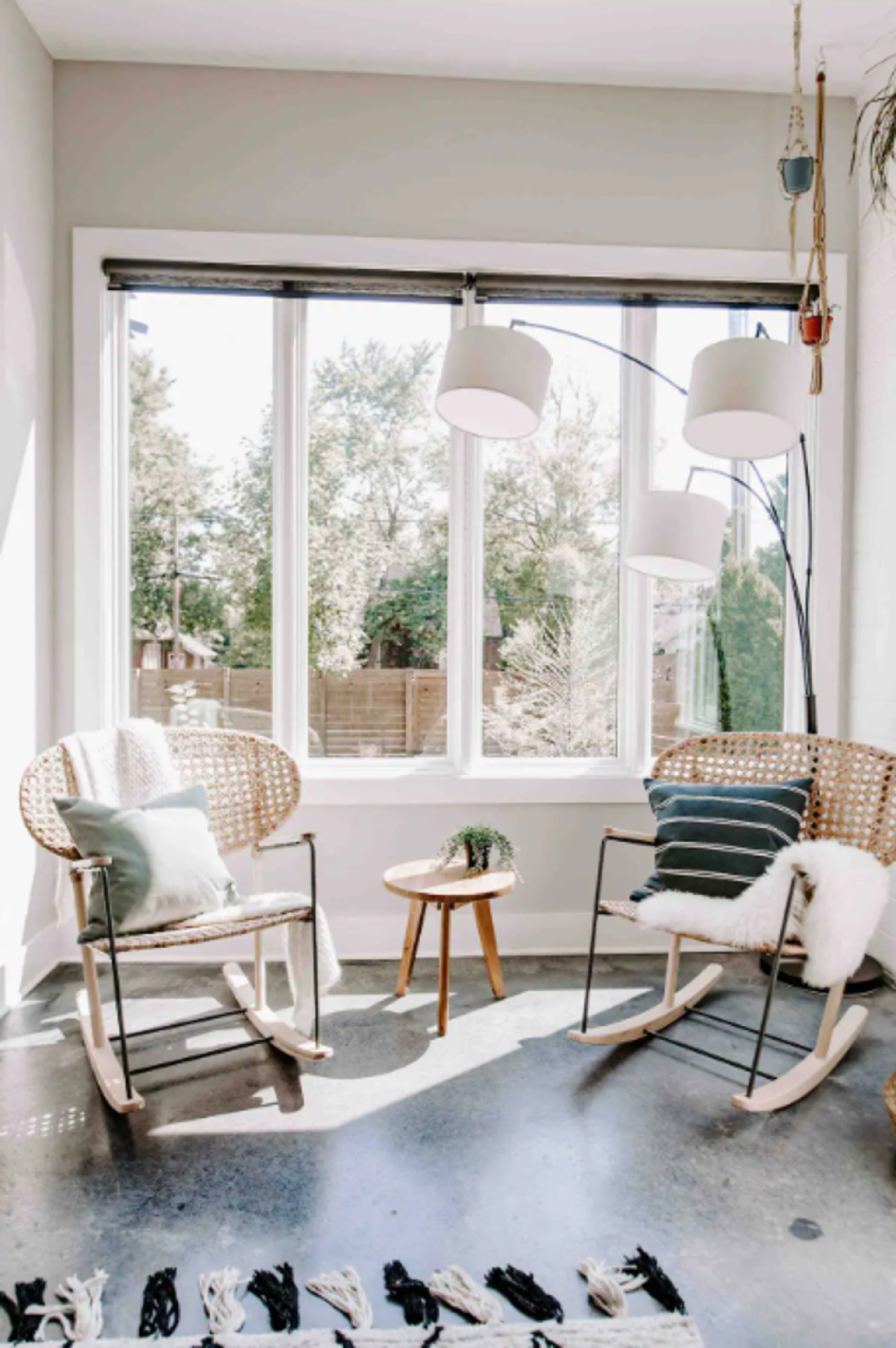 Two rocking chairs with cushions are positioned next to a small table in a sunlit corner with large windows.
