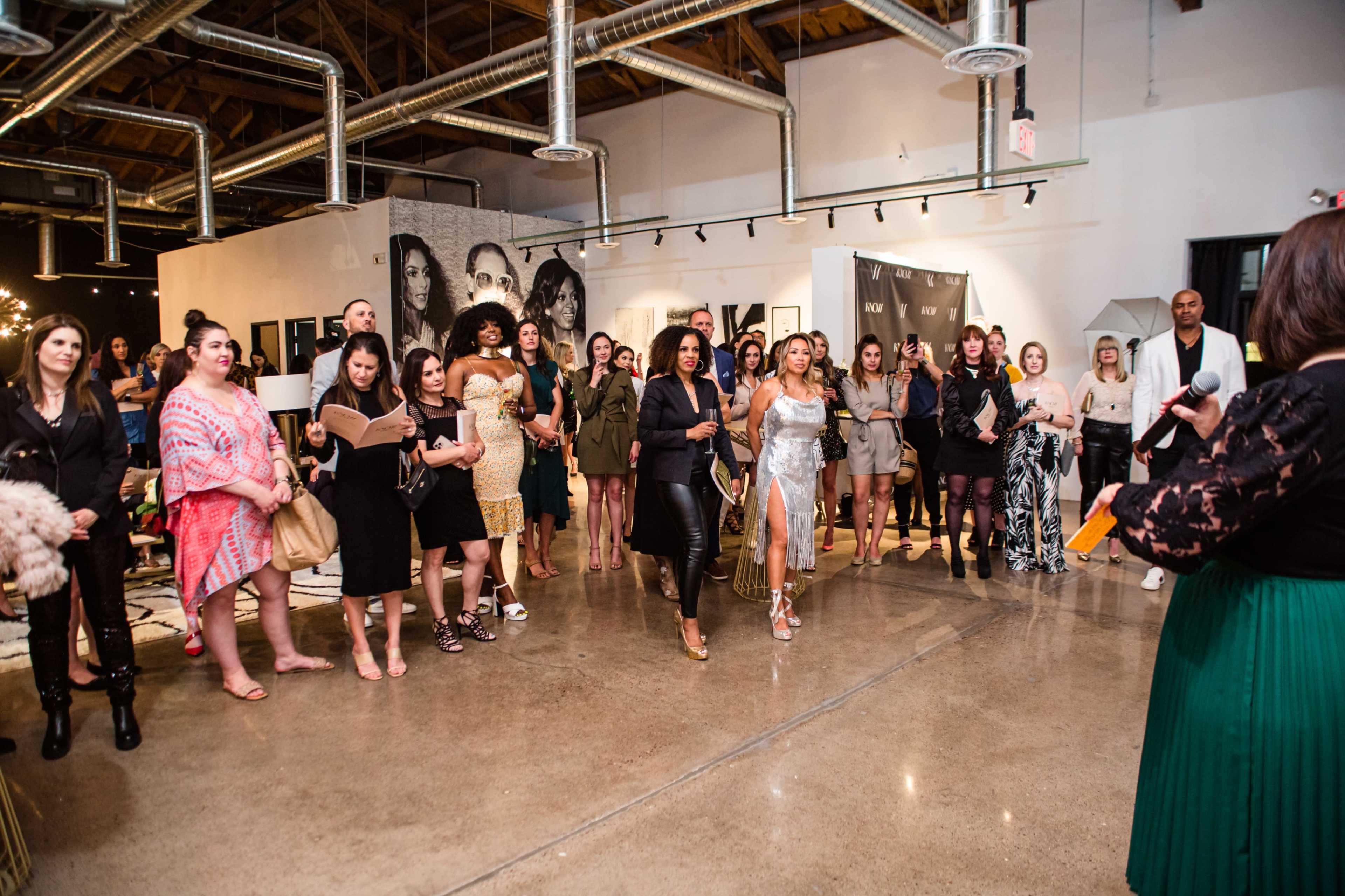 A group of women attends an event in a spacious gallery, while one woman in a silver dress walks towards the front.