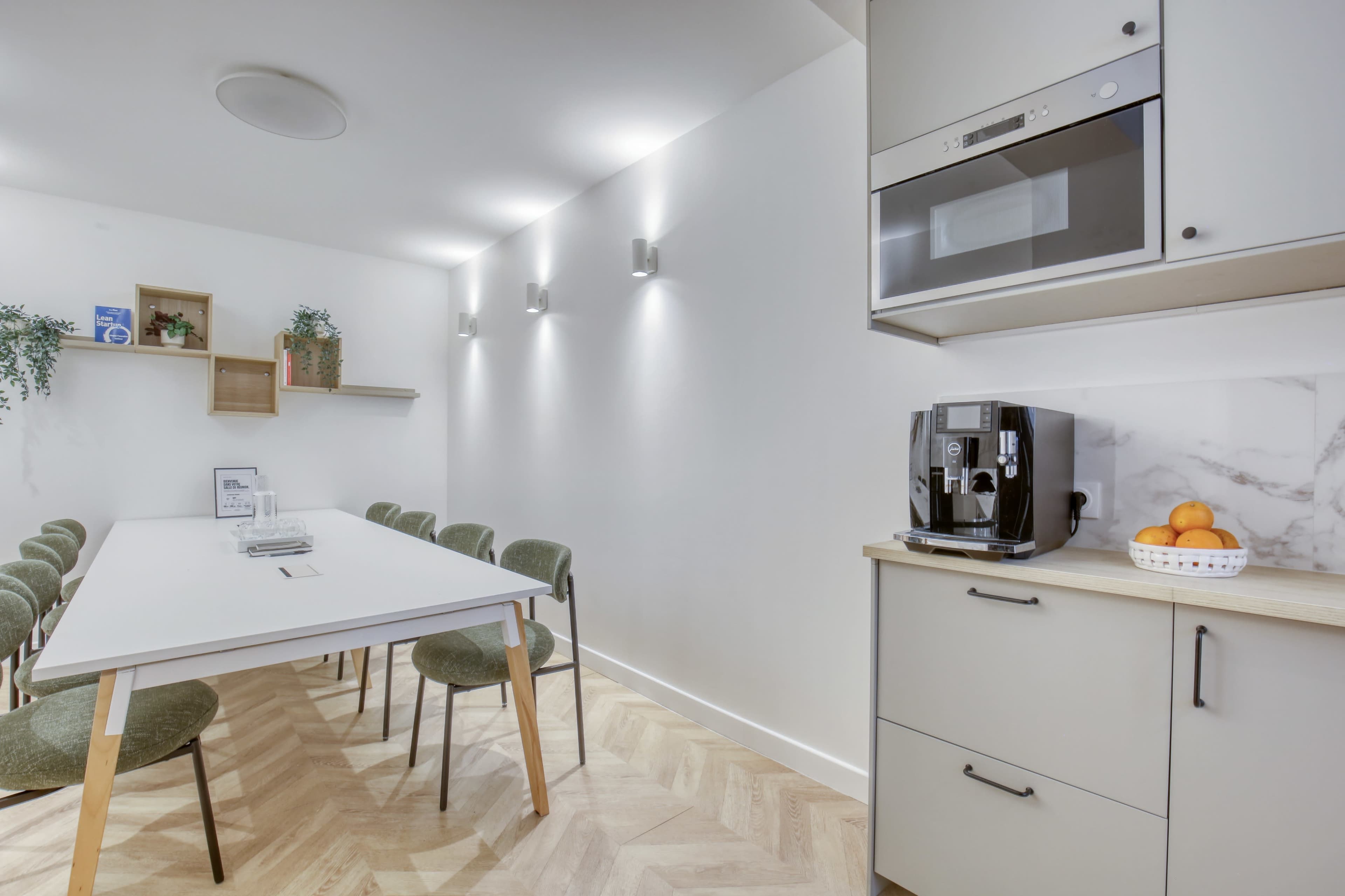 The image shows a modern kitchen space featuring a long dining table with green chairs, a wall-mounted microwave, and a coffee machine, set against a minimalist white background with decorative plants.