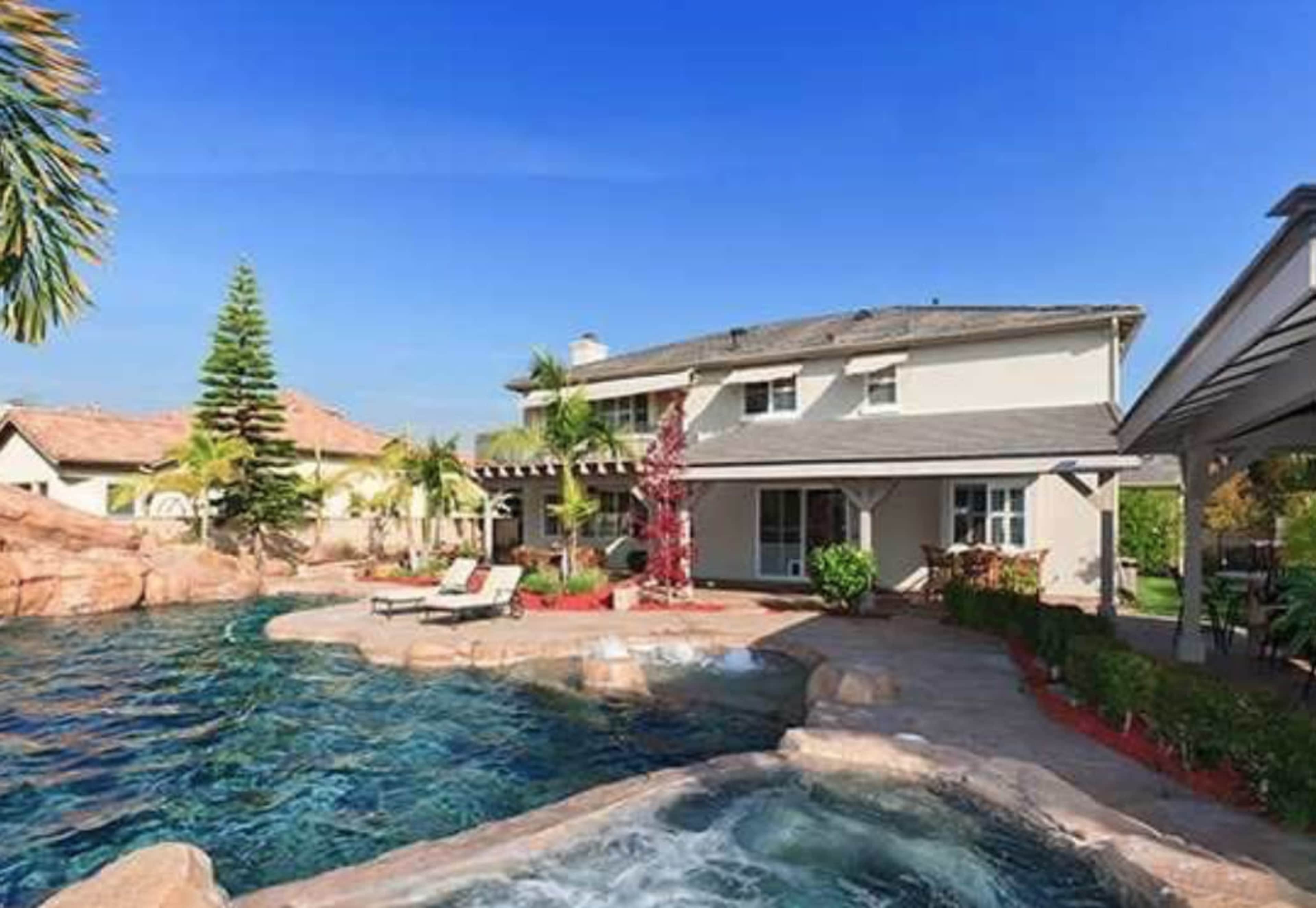 The image depicts a spacious backyard featuring a swimming pool, stone landscaping, and a two-story house with a patio area.