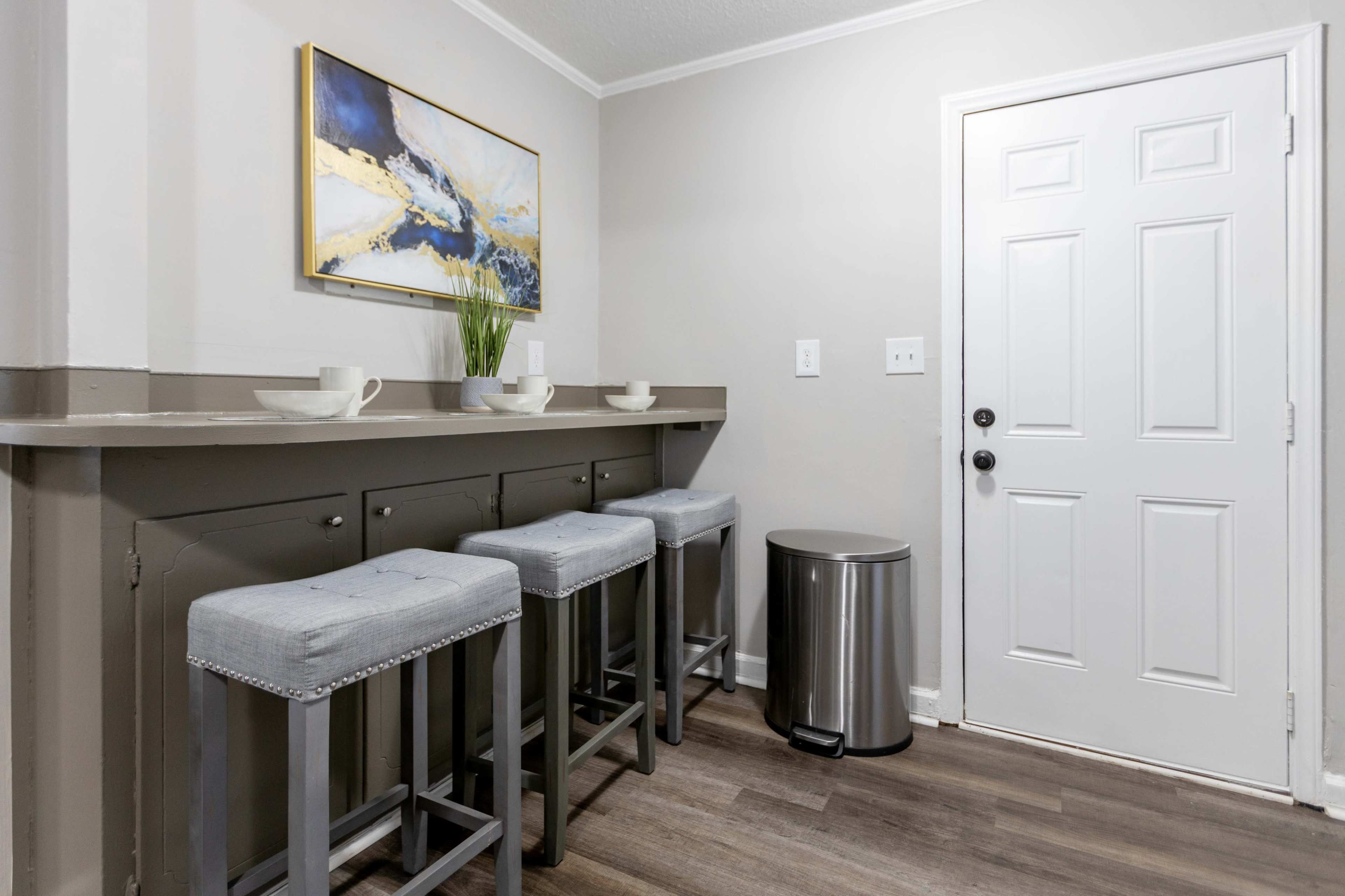 A narrow space features four bar stools along a low counter, a small table with cups, and a door leading outside next to a stainless steel trash can.