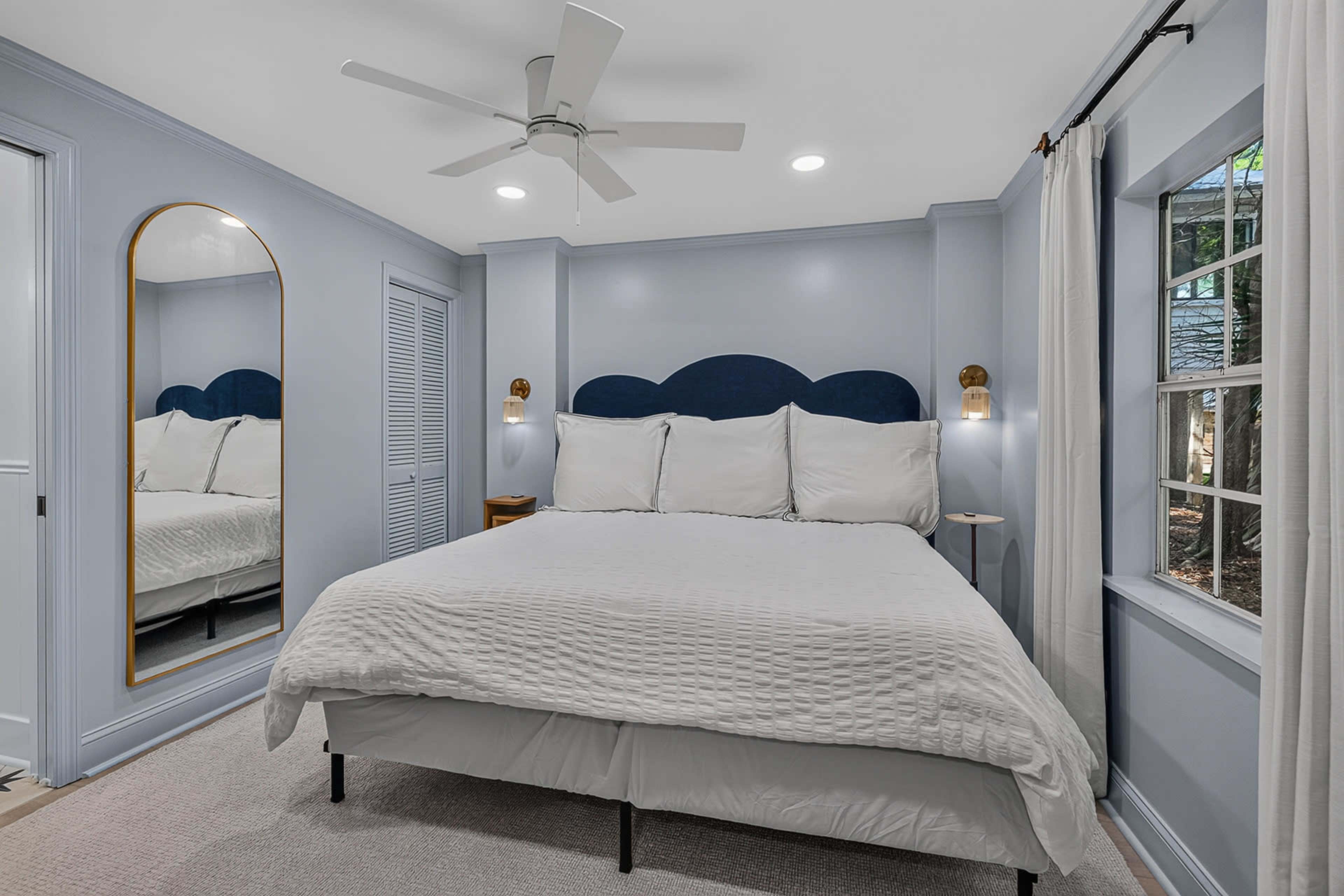 A bedroom features a large bed with white bedding, a blue upholstered headboard, and a small nightstand, all set against light blue walls and complemented by natural light from the windows.
