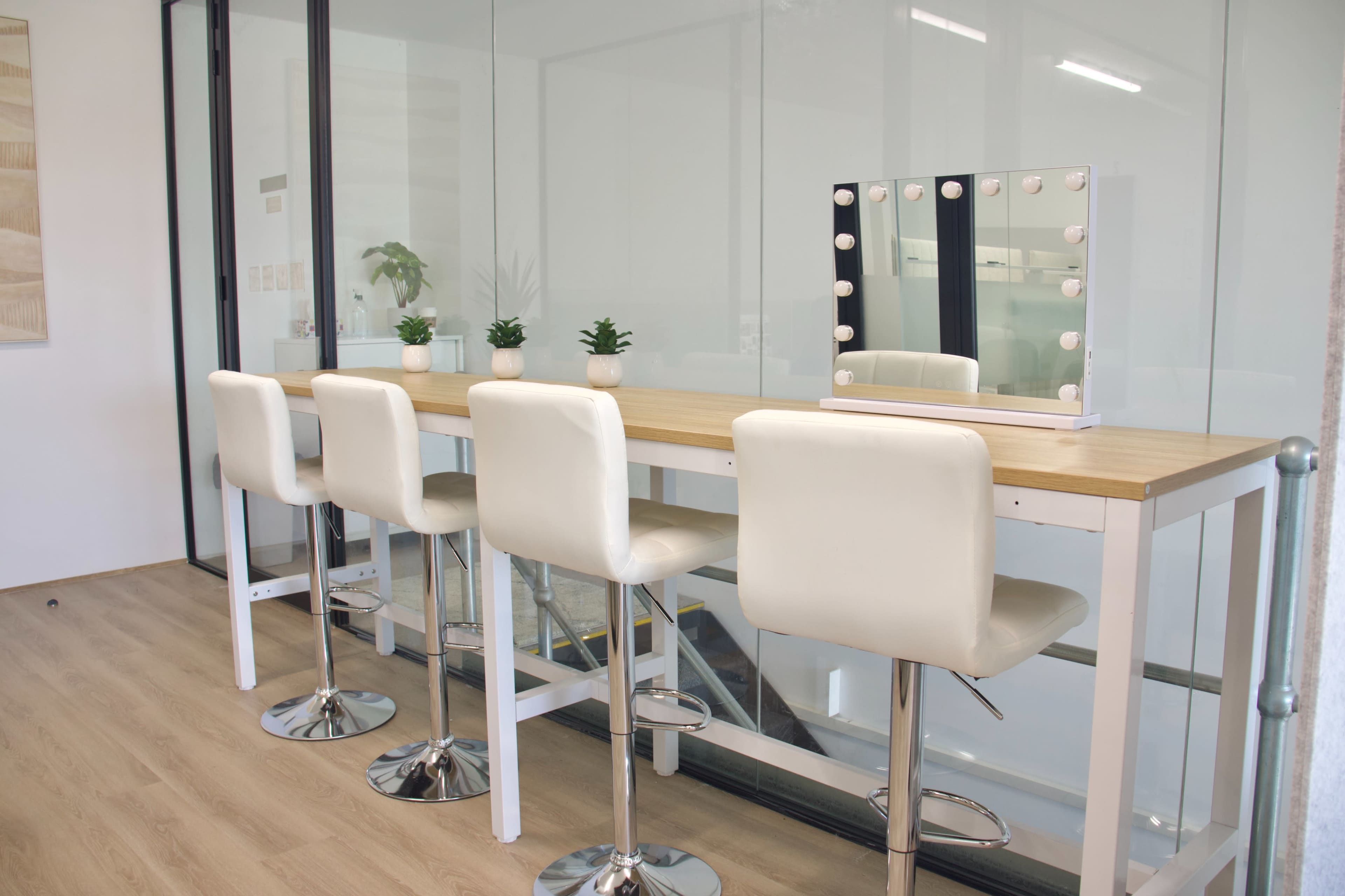 A sleek vanity area features a long wooden table with four white bar stools and a mirror illuminated by bulbs.