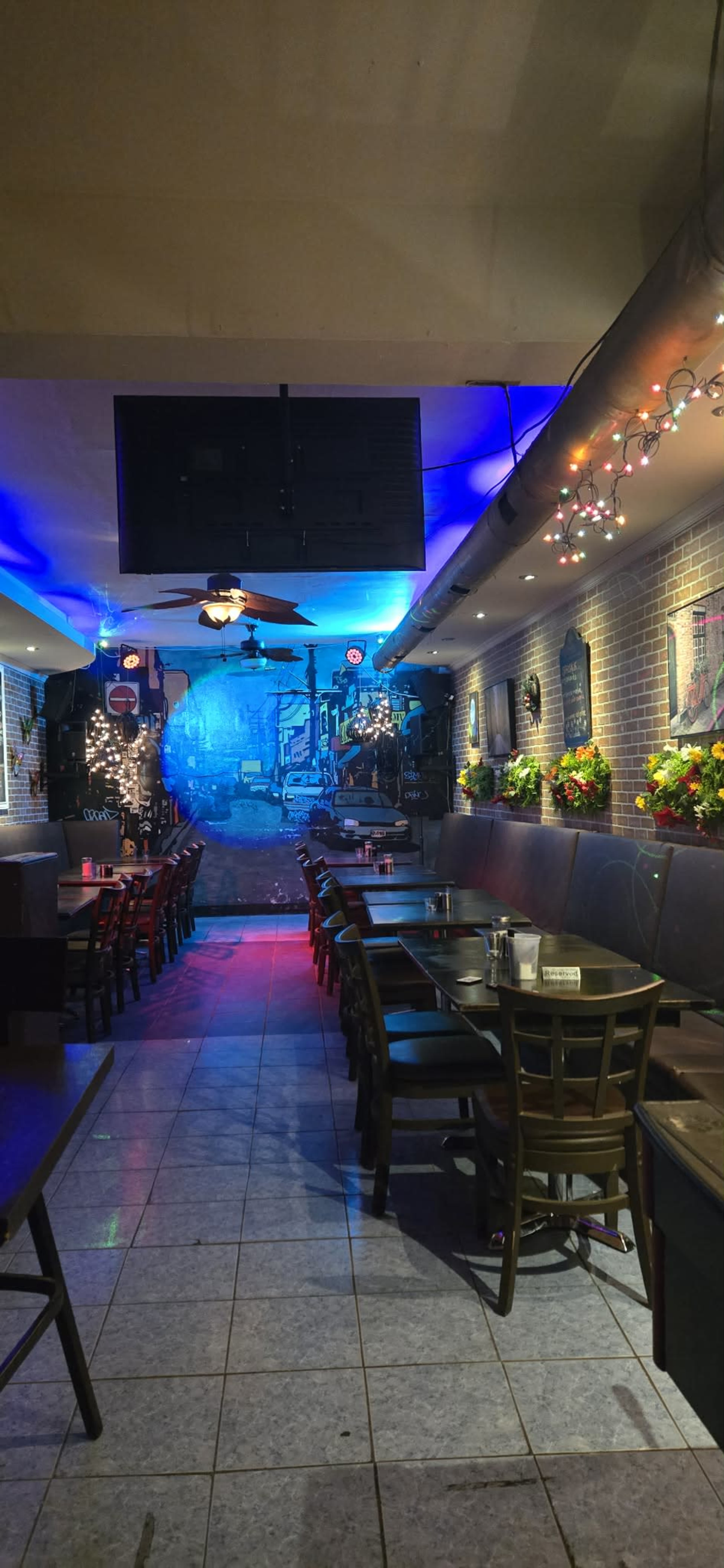 The image shows the interior of a restaurant featuring tables and chairs arranged in a narrow layout, with a mural depicting a street scene on the back wall and colorful lights enhancing the ambiance.
