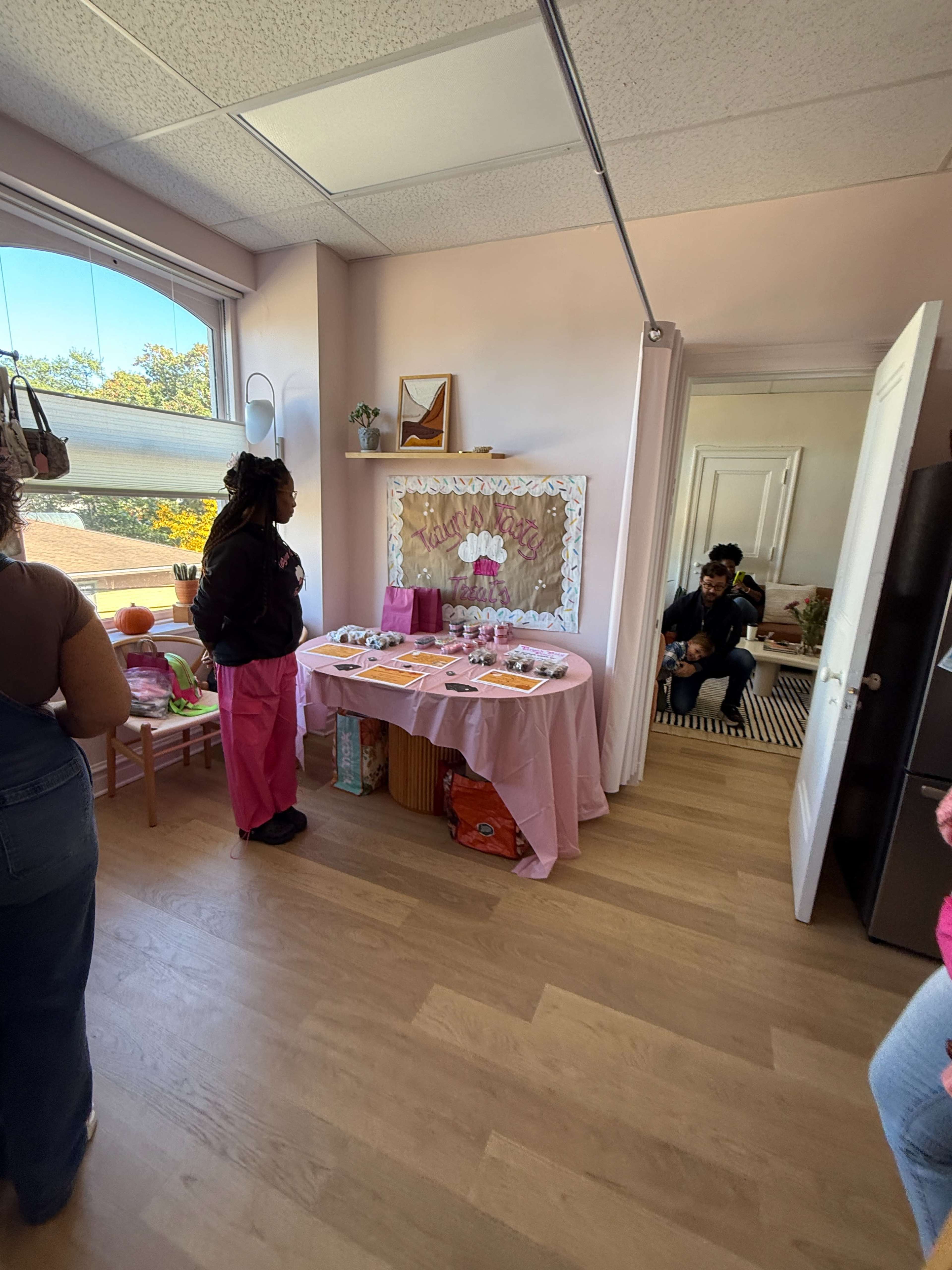 The image shows a decorated room with a table covered in pink cloth and various items displayed on it, while several people engage in conversation.