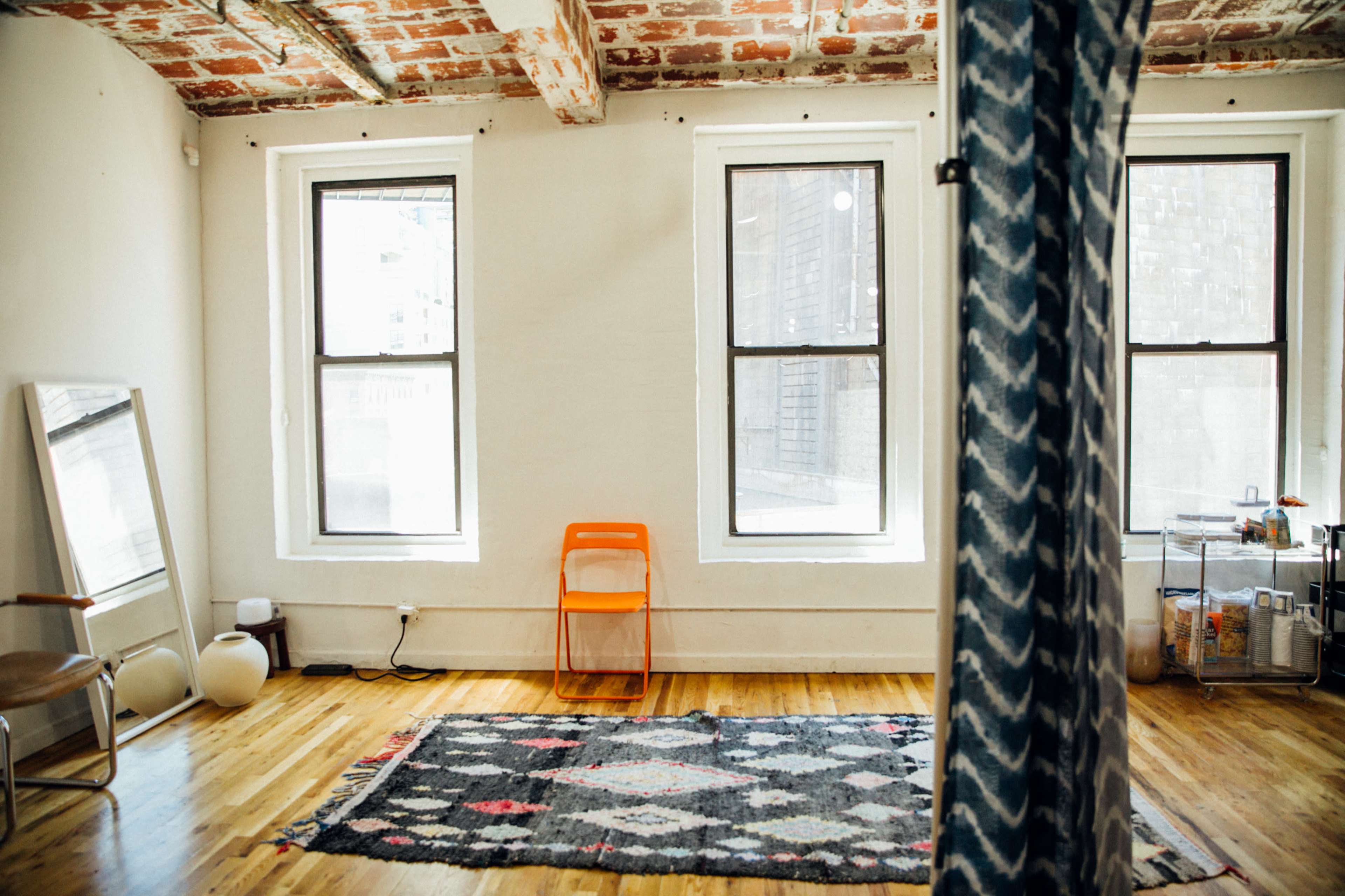 A minimalist room with wooden floors, three windows, an orange chair, a patterned rug, and a mirror against the wall.