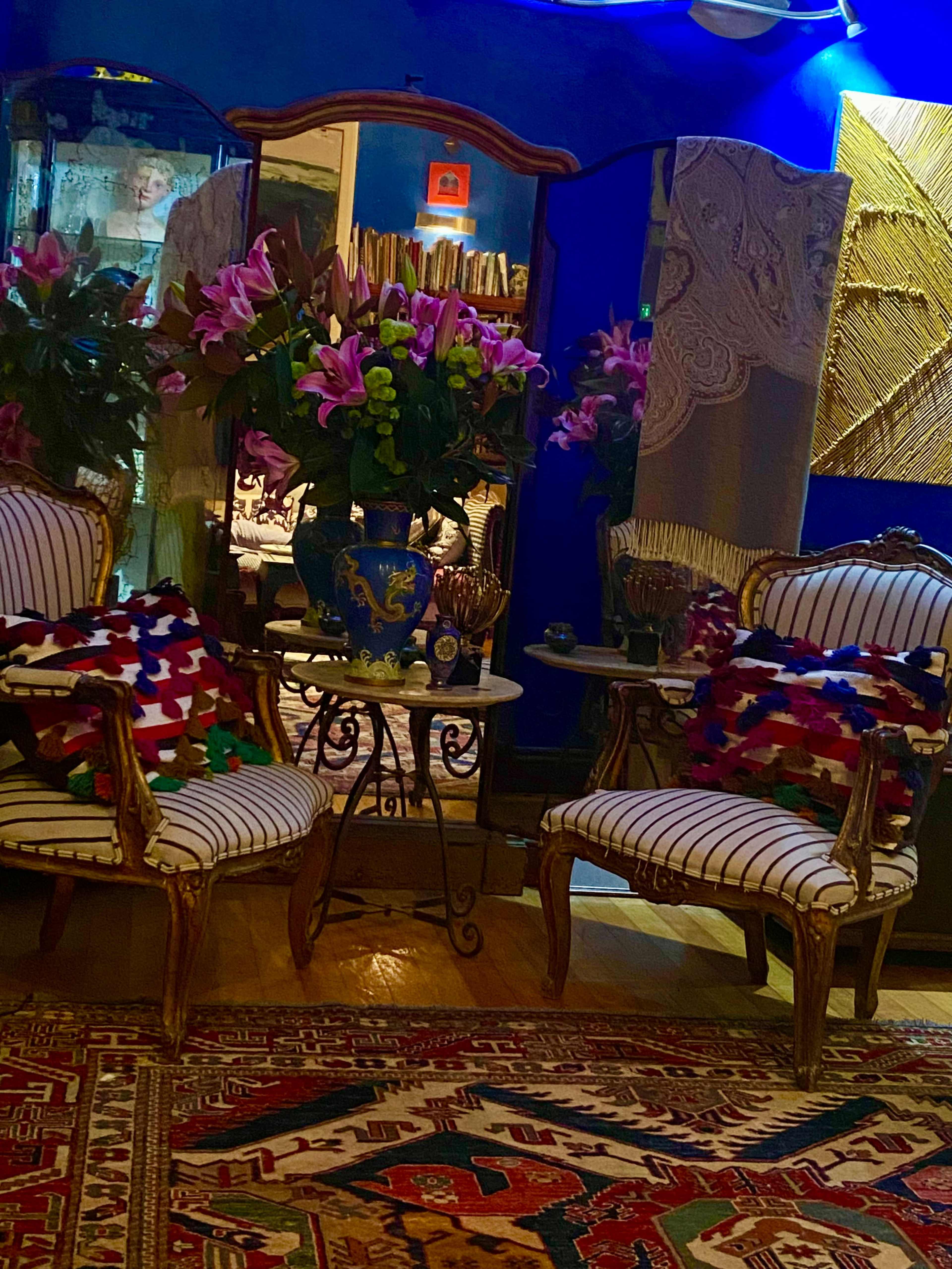 The image depicts an elegantly styled room featuring two ornate chairs facing a small round table, adorned with vibrant flowers and a colorful area rug.