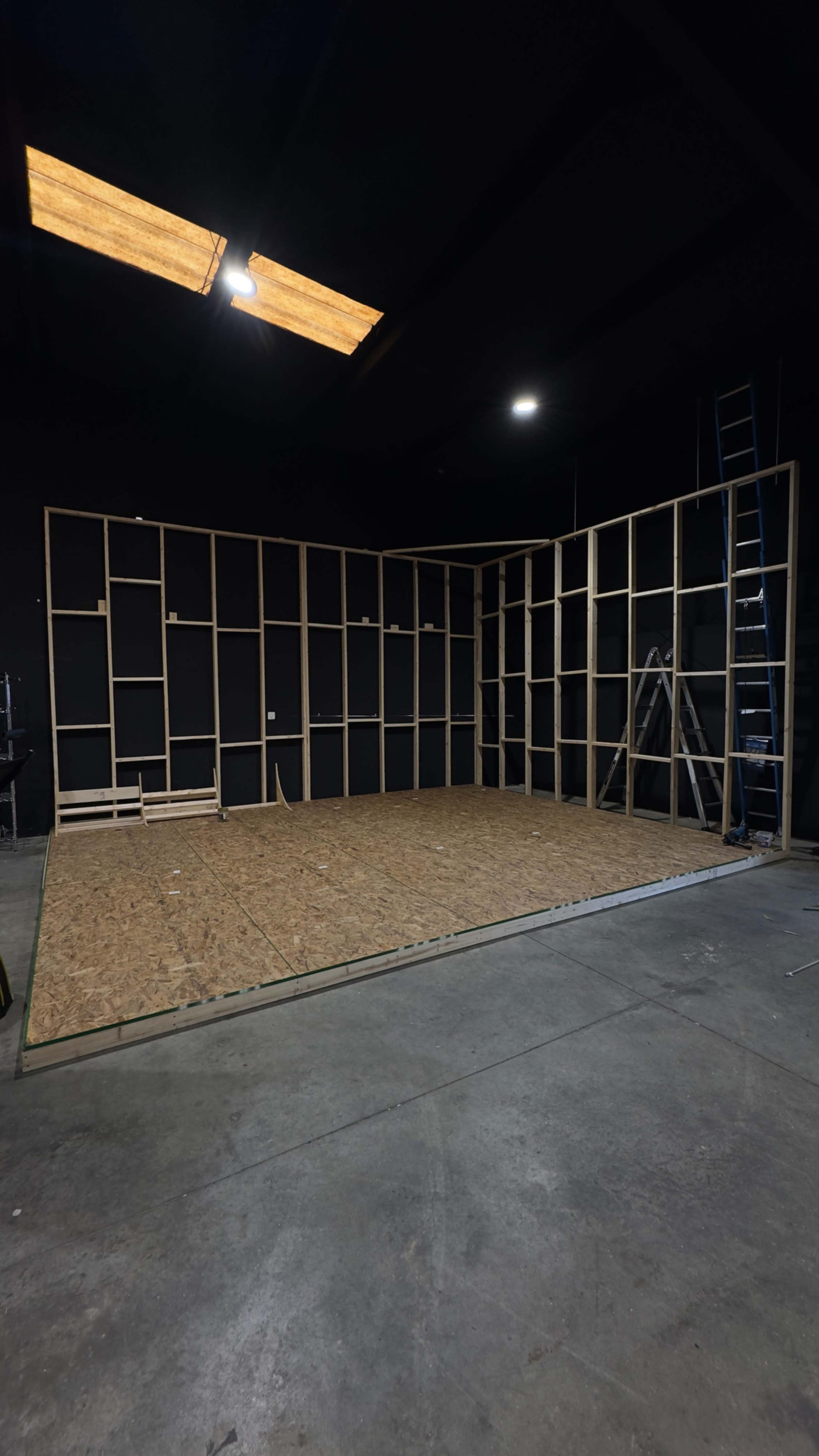 A large, empty room features a wooden frame structure against the walls and a plywood floor, with a ladder positioned nearby.