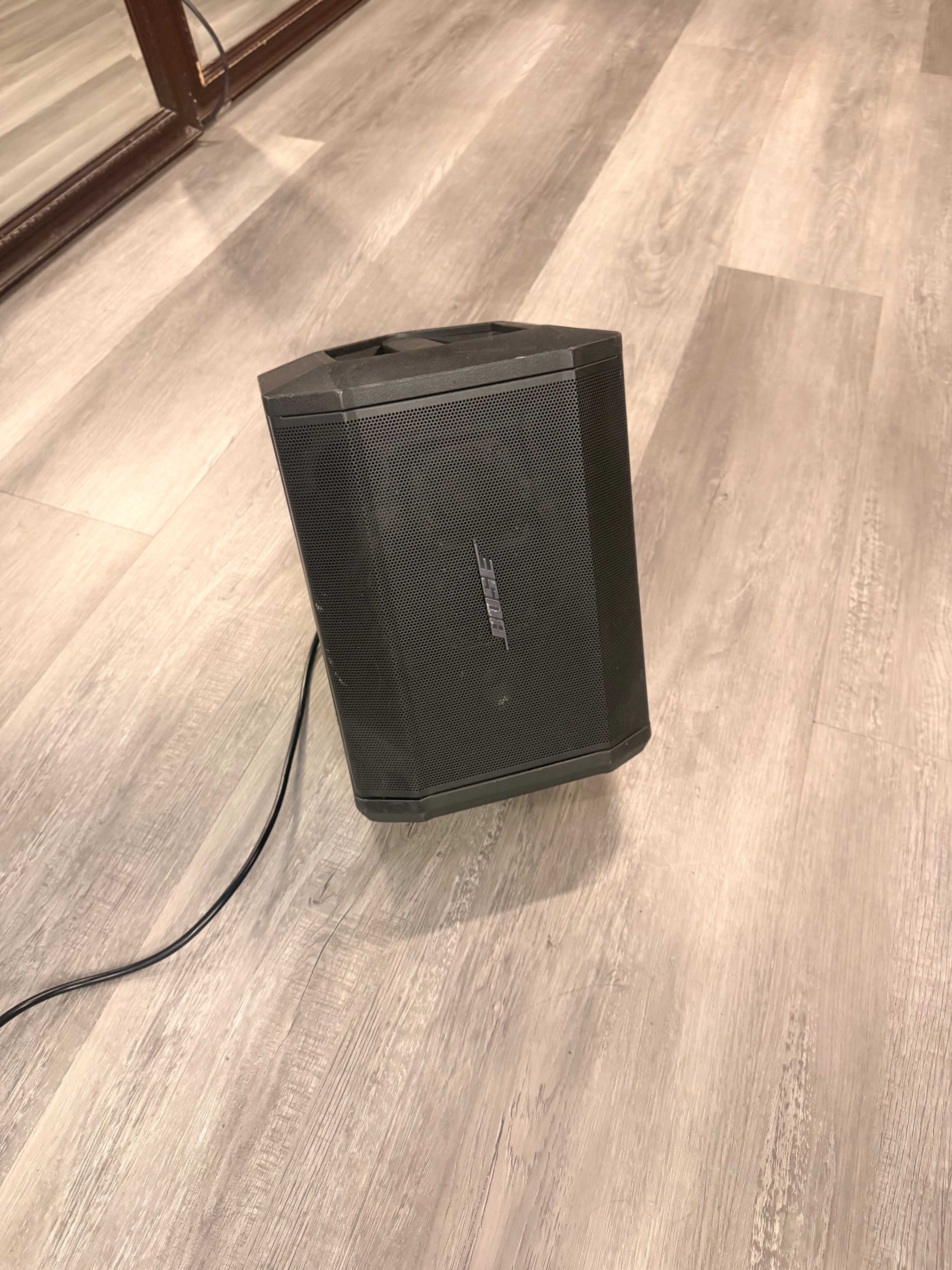 A black Bose speaker is positioned on a light-colored wooden floor.
