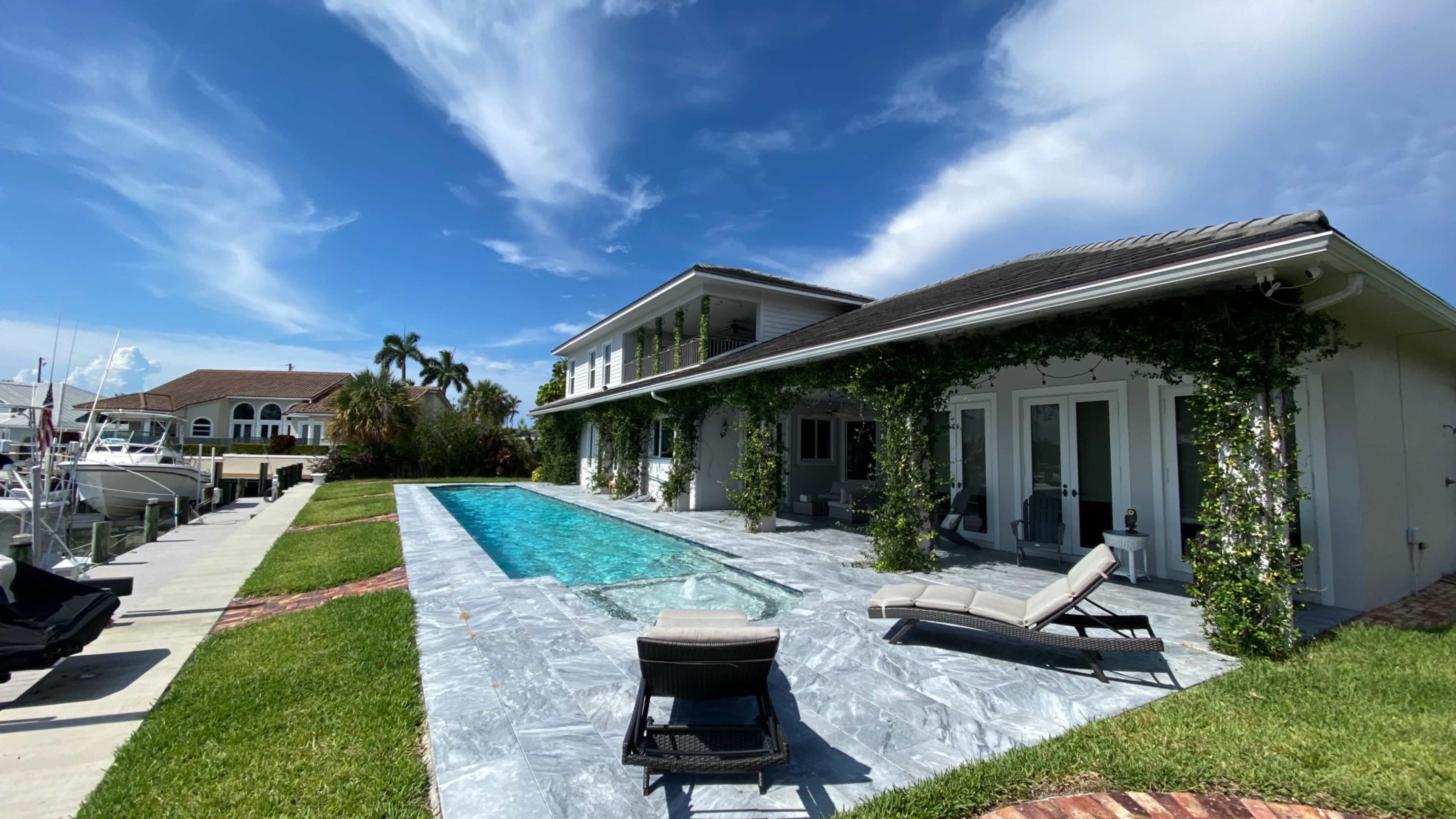 A modern house with a swimming pool and lounge chairs, situated beside a marina with boats in the background.