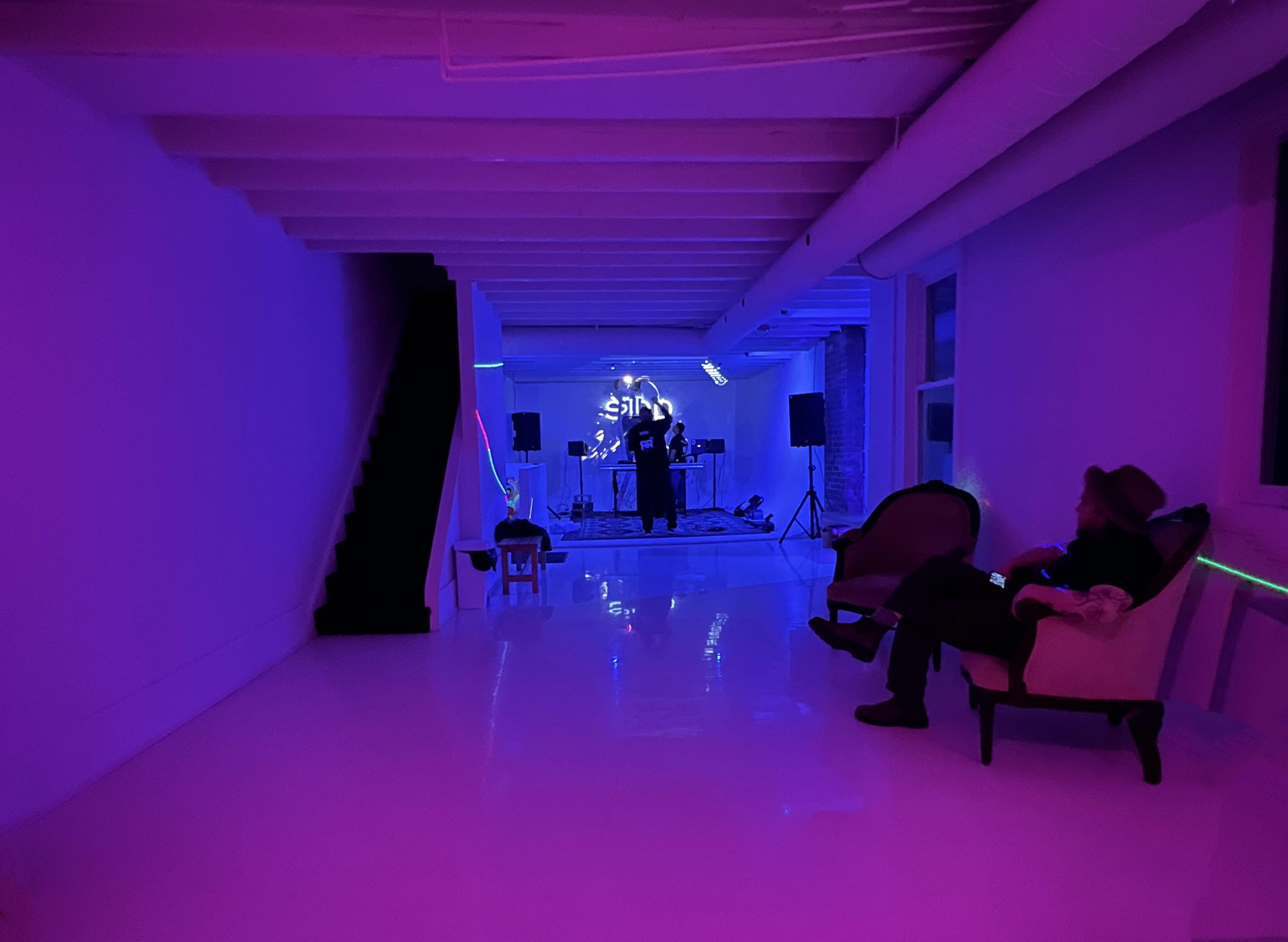 A musician performs on stage in a dimly lit room with purple and blue lighting, while a person sits on a couch in the foreground.