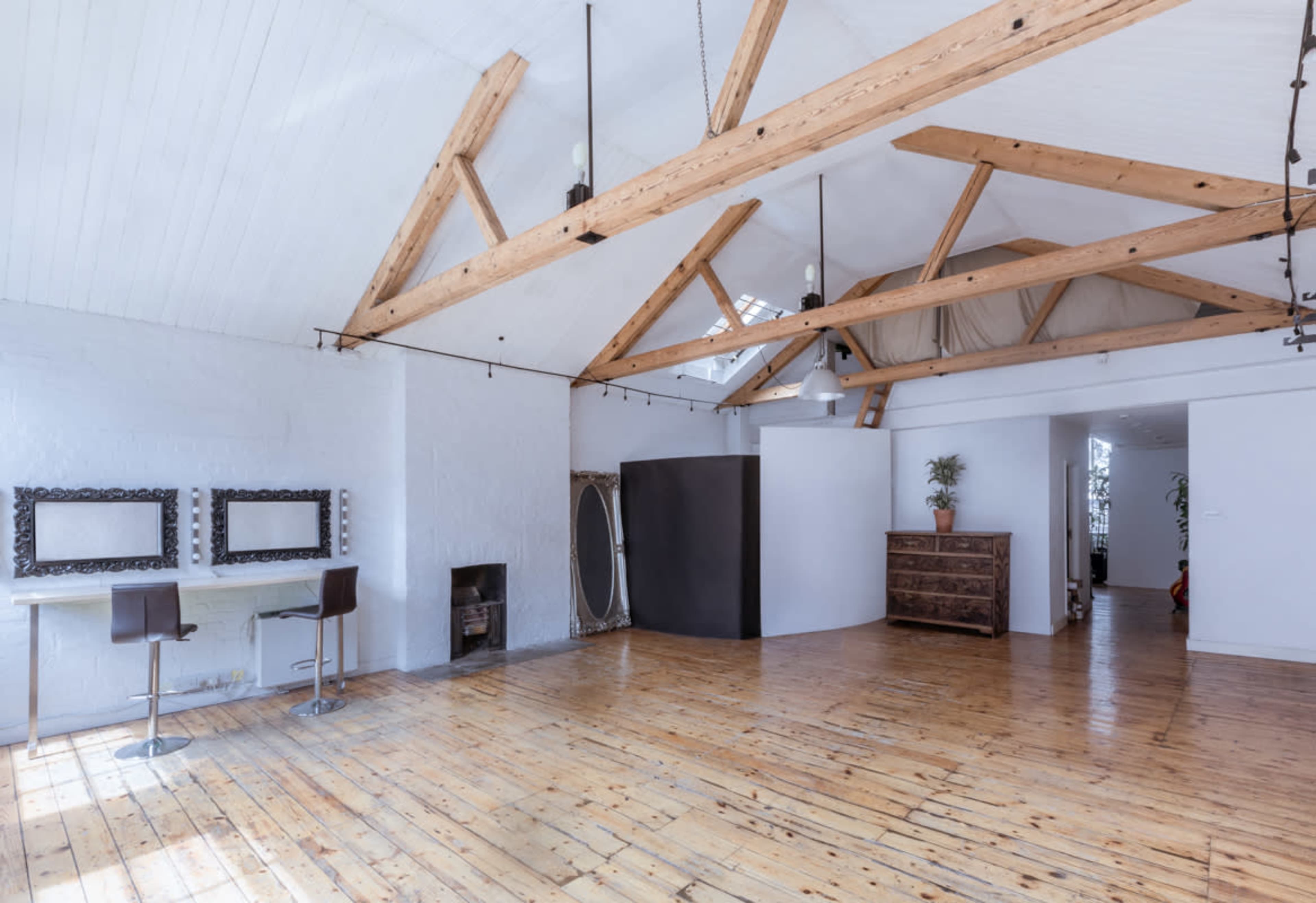 A spacious, bright room with wooden floors, exposed beam ceilings, and a minimalist layout featuring a desk, two chairs, a mirror, and a dresser.
