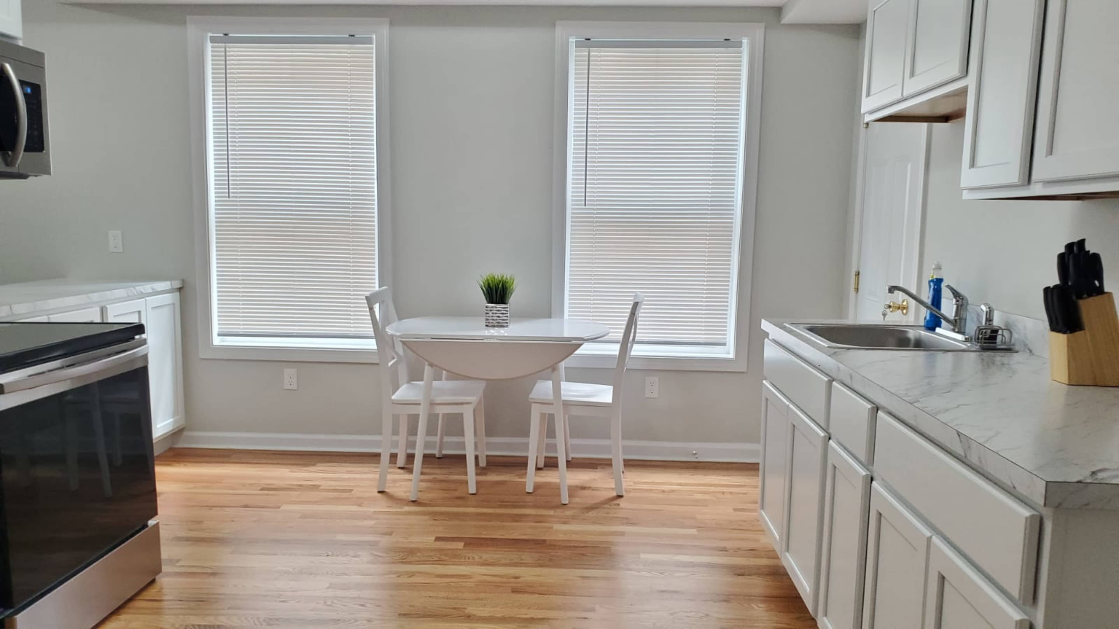 The image shows a simple kitchen with a round table and two chairs beside large windows, featuring wooden floors and white cabinetry.