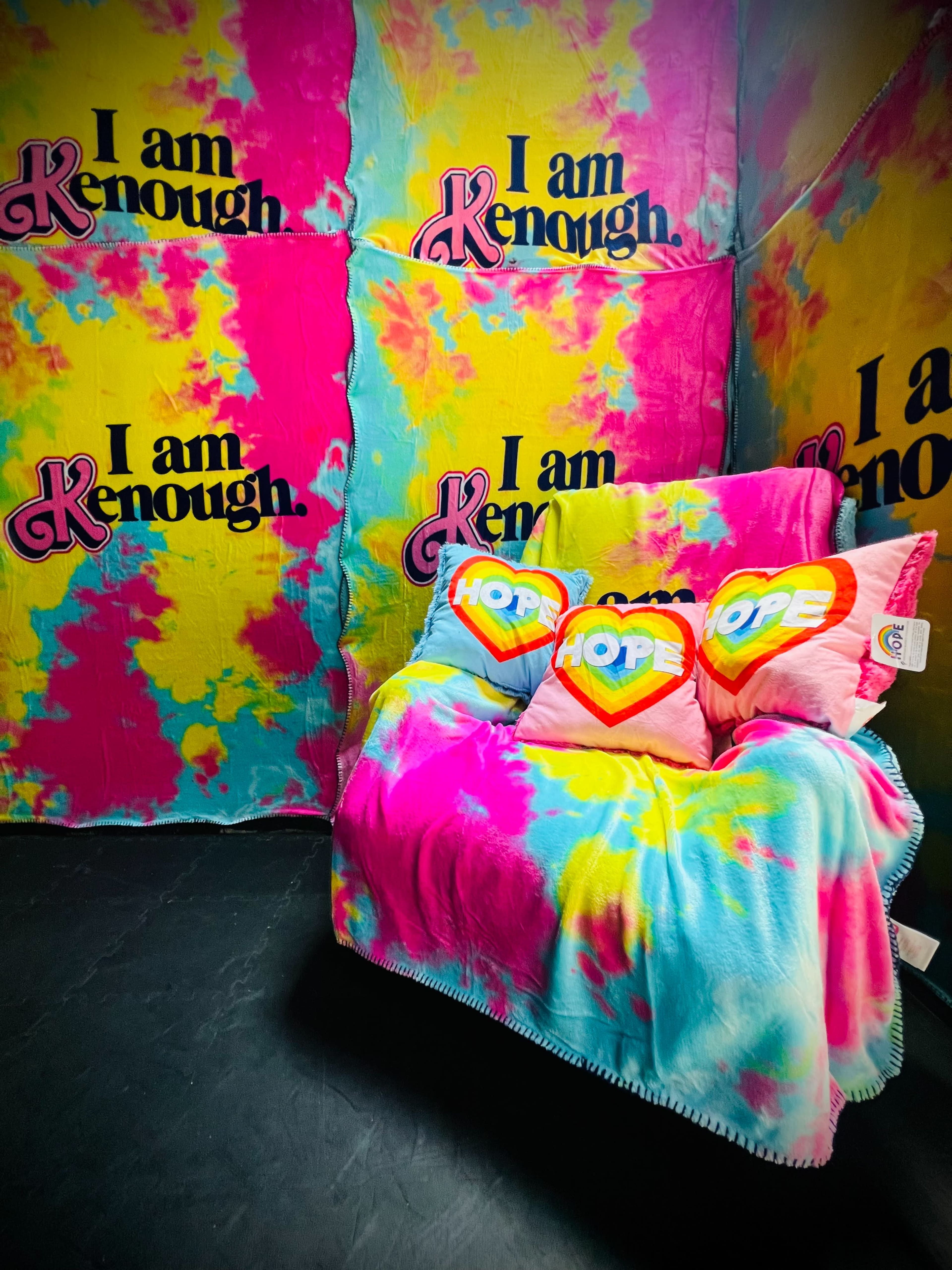 The scene features a brightly colored tie-dye backdrop with the phrases "I am Kenough" and decorative pillows that read "HOPE" on a cozy couch.